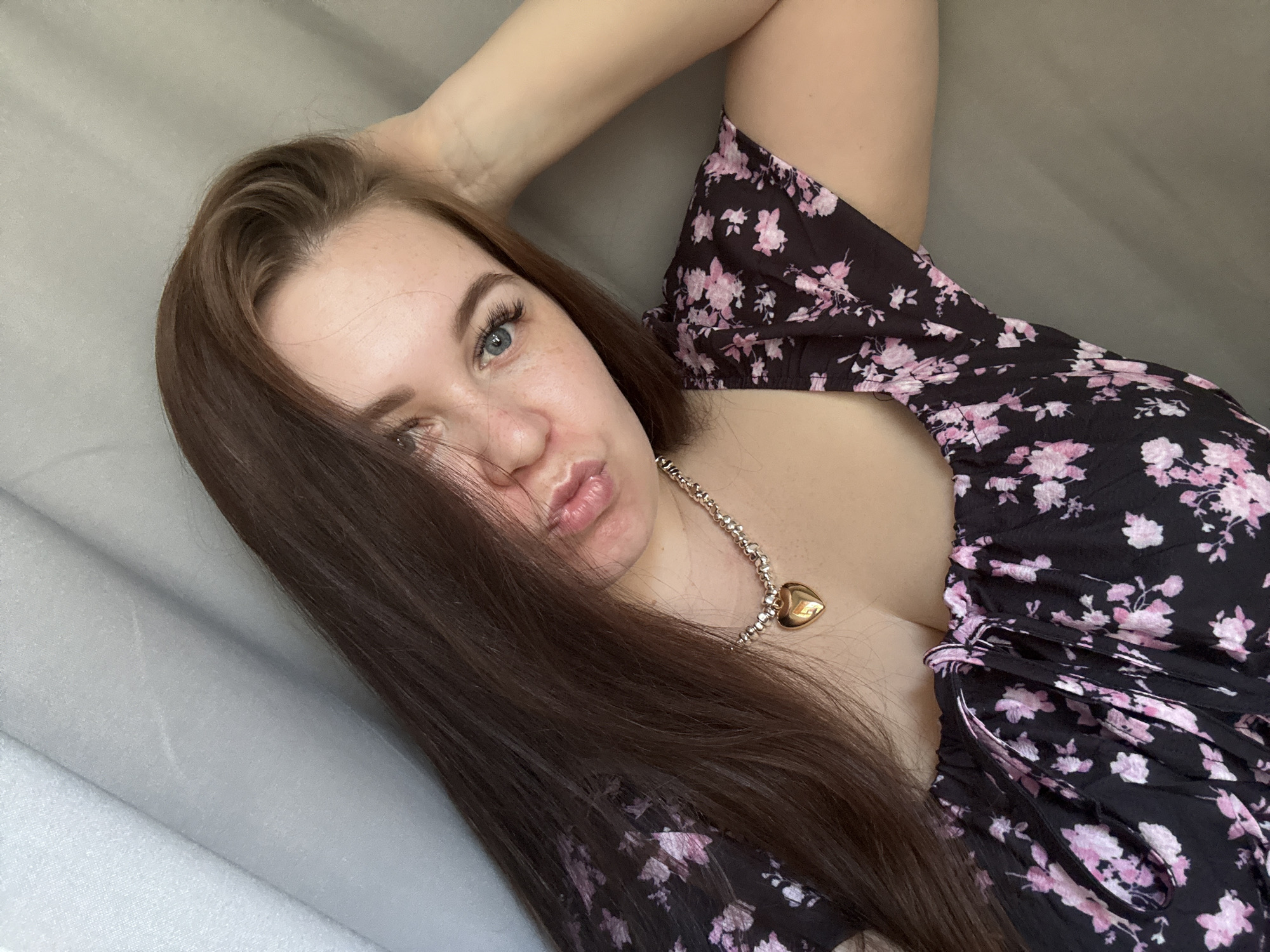 A young woman with long, dark brown hair and blue eyes is pictured lying down, looking at the camera. Her hair is partially covering her face. She is wearing a black dress with a pink floral pattern and a silver necklace with a heart-shaped pendant. The woman has her arm behind her head and her lips are slightly pursed. Her facial expression could be interpreted as playful or sultry.
The setting appears to be indoors, with a soft, grey background that suggests a bed or a sofa. The lighting is soft and diffused, indicating it might be daytime. There is no visible text in the image. The overall mood is casual and intimate.