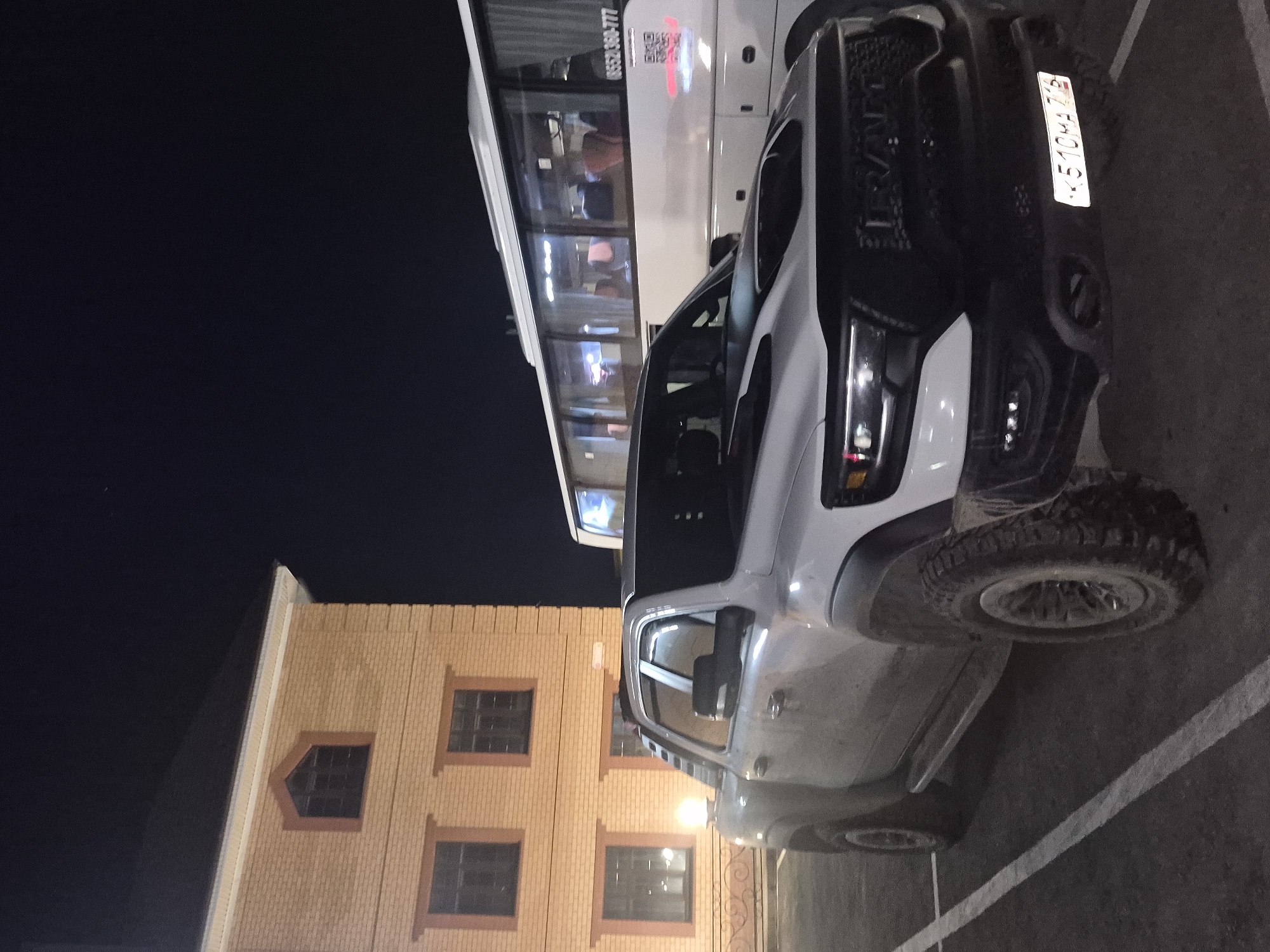 At night in Sergeevka, Russia, a white Ram TRX pickup truck with Russian license plate К 510 ЧА 75 is parked next to a white bus with passengers. A two-story building with brick facade and several windows is visible behind the truck. The scene is in a parking lot marked with white lines.