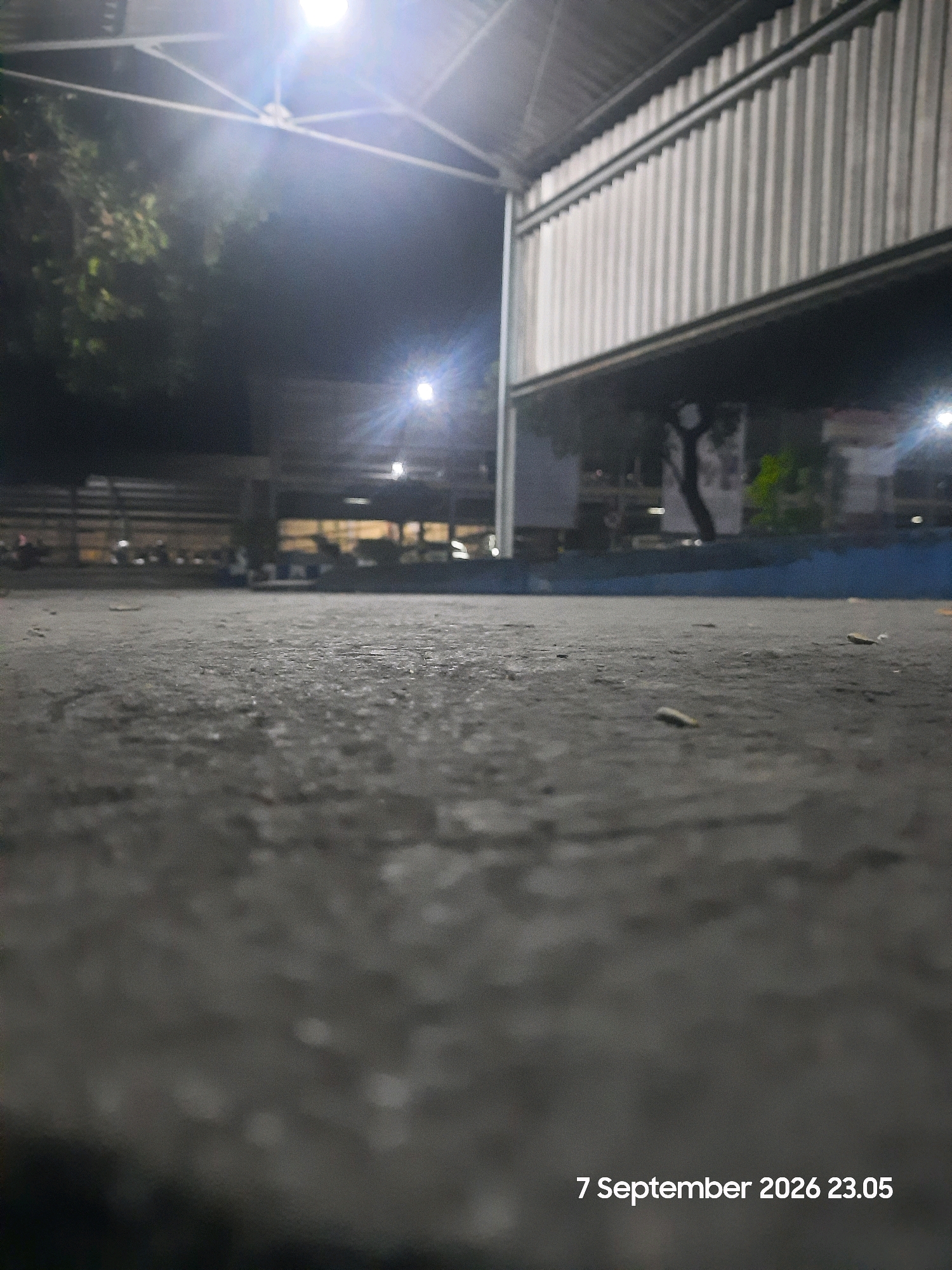 The image depicts a low-angle, nighttime view of a paved outdoor area, possibly a sports court or a public gathering space, located in Gending, Indonesia. The foreground is dominated by a textured, grey concrete surface, with a few small, indistinct objects scattered on it. In the mid-ground and background, the scene is dimly lit by artificial lights.
To the left, trees with foliage are visible, partially obscured by shadow and the bright glare of overhead lighting. A structure with a corrugated metal roof and supporting beams is prominent on the right, casting shadows onto the ground. Further back, there are indistinct structures and what appear to be parked vehicles or motorbikes. The overall environment suggests an open-air venue at night, likely with some form of roof or canopy overhead. The date and time "7 September 2026 23.05" are superimposed on the bottom right corner of the image.