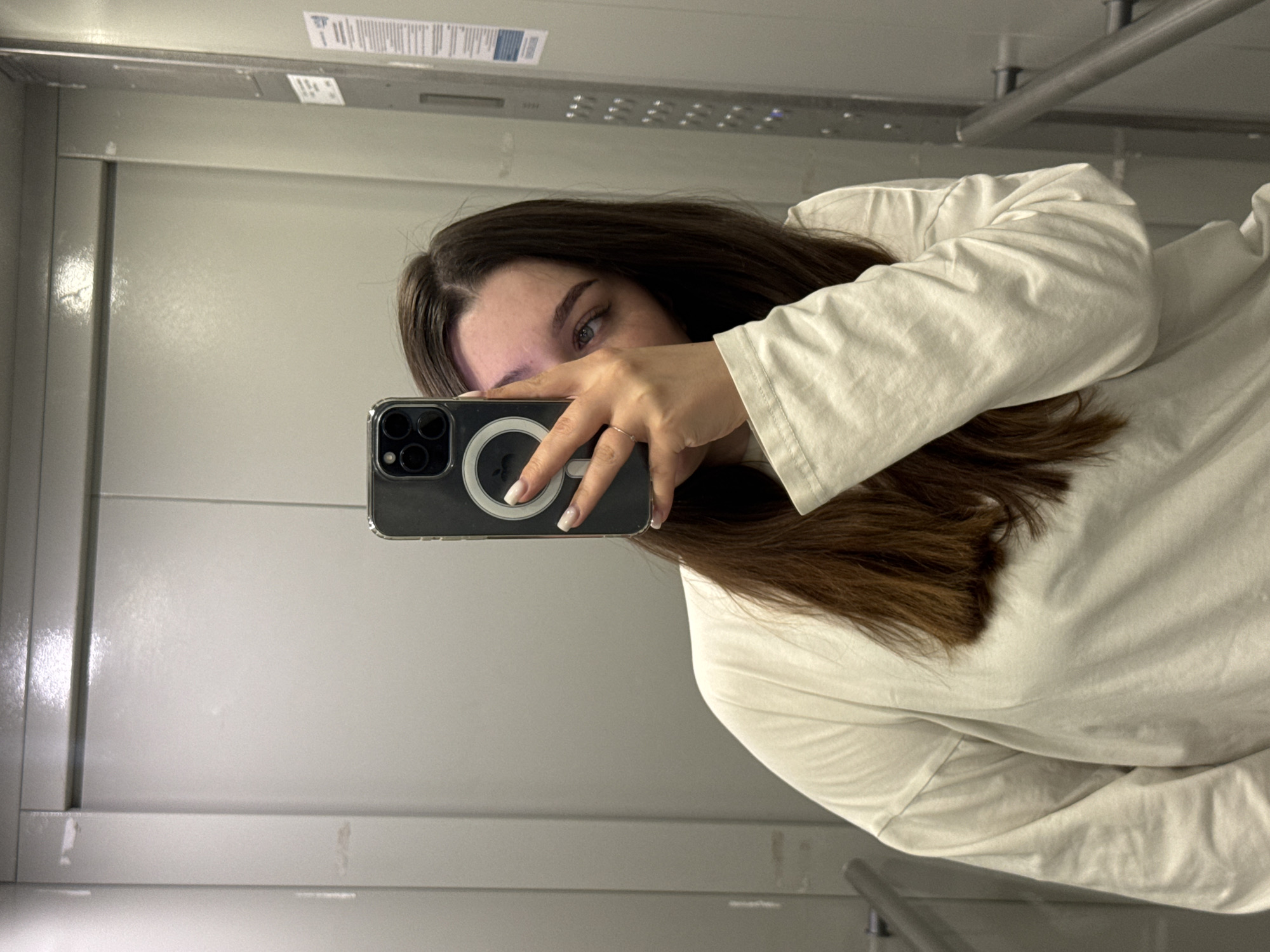 A woman stands in what appears to be an elevator, taking a selfie in the mirror with a modern smartphone. The phone has a case with a magnetic ring on the back. Only a portion of her face is visible behind the phone; the woman has brown hair. She is wearing a white long-sleeved shirt. The elevator's interior is metallic gray. A handrail is visible in the upper right. There is nothing in the image to indicate this is Dikaya Gar', Russia.