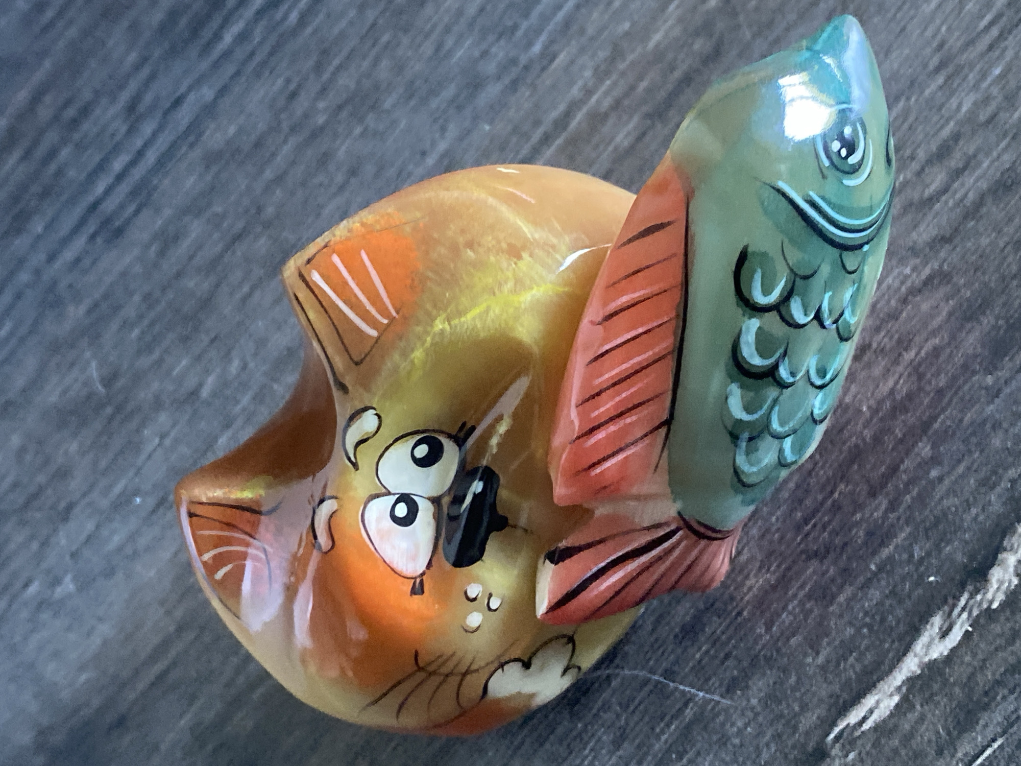 A decorative ceramic figurine of a cartoon cat and fish lies on a dark, wooden surface. The cat is depicted in shades of orange and yellow, with wide, surprised eyes, a black nose, and white whiskers. It has orange stripes on its ears. A red-tailed fish, with green scales and blue accents, is positioned in front of the cat. The fish has a single eye and appears to be the object of the cat's attention. The wooden surface has a visible grain pattern and appears to be a tabletop or floor. The lighting suggests an indoor environment. The location of Novokuznetsk, Russia is not discernible from the image itself.