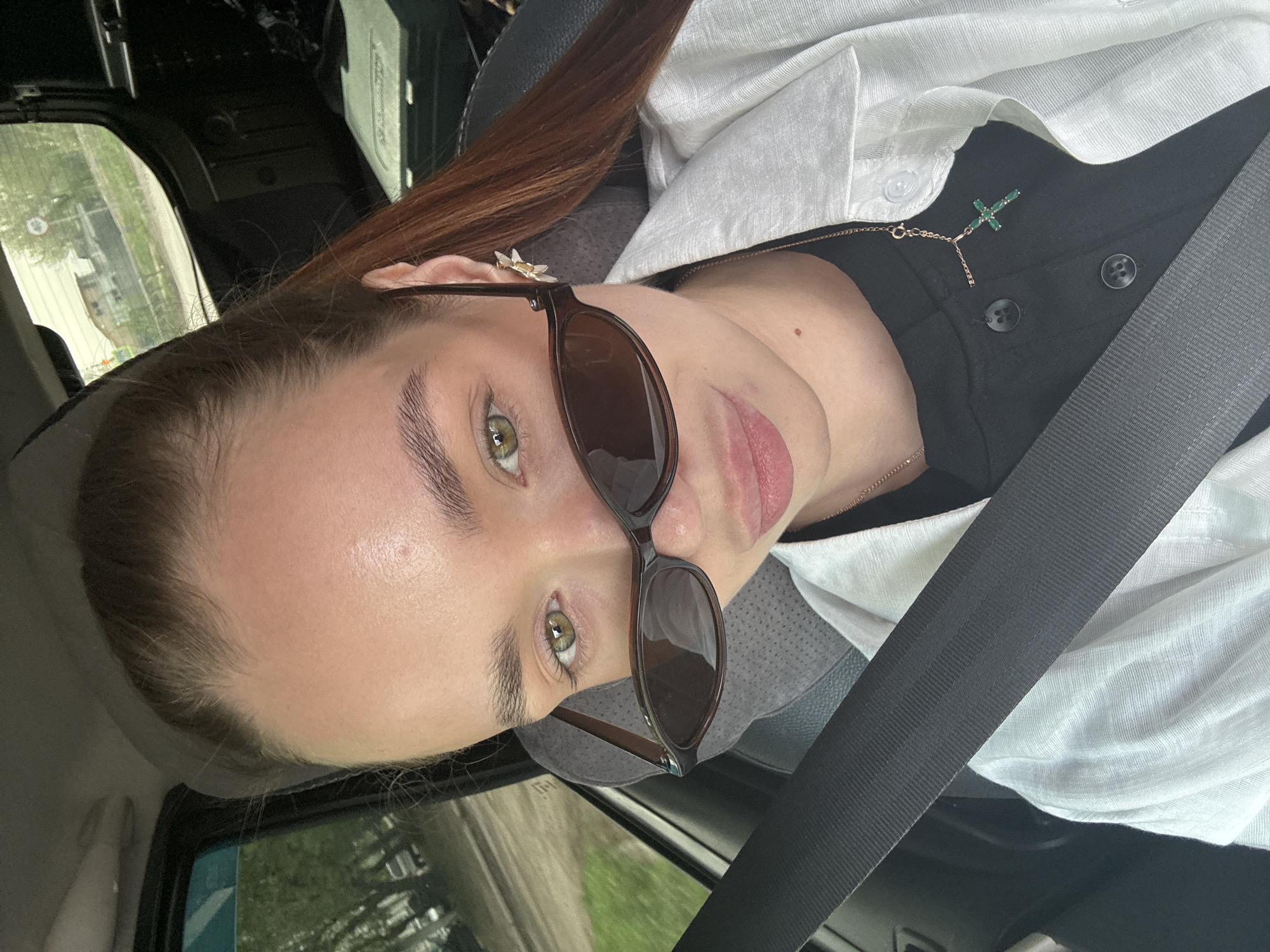 A woman is in the driver's seat of a car, wearing sunglasses and a seatbelt. She has light skin, brown hair, and green eyes. She is wearing a black shirt under a white button-down shirt. A necklace with a green cross pendant is visible. Outside the car window, a blurred street scene is visible, suggesting a city environment. This cannot be confirmed to be in Chaikovsky, Russia, based on the provided image.