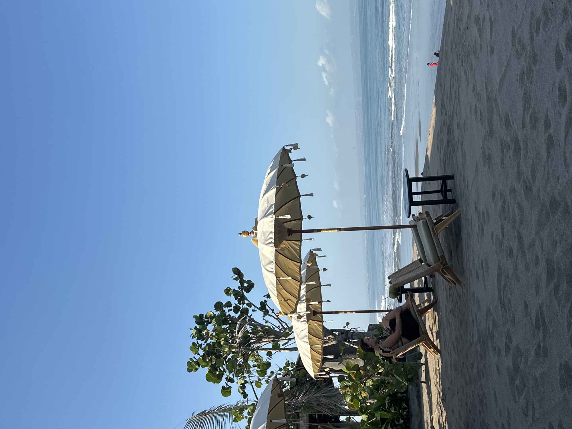 This image depicts a serene beach scene in North Kuta, Indonesia. In the foreground, dark grey sand is textured with subtle footprints and ripples. A woman, wearing a black and white patterned top and black bottoms, is seated in a wooden beach chair on the sand, facing the ocean and holding a coconut drink. To her right, another identical empty beach chair is positioned. A small, dark, round-topped table stands between the chairs.
Behind the seating area, two large, light-colored parasols with tassels provide shade. Lush green foliage and trees are visible to the left of the parasols, suggesting a vegetated area adjacent to the beach. The ocean stretches out to the horizon under a clear blue sky, with gentle waves lapping the shore. In the distance, faint clouds are present near the horizon line. Further down the beach, two small figures can be seen, one wearing red and the other a dark color, possibly a child and a dog. The overall atmosphere is tranquil and relaxed, indicative of a beach resort or cafe setting.