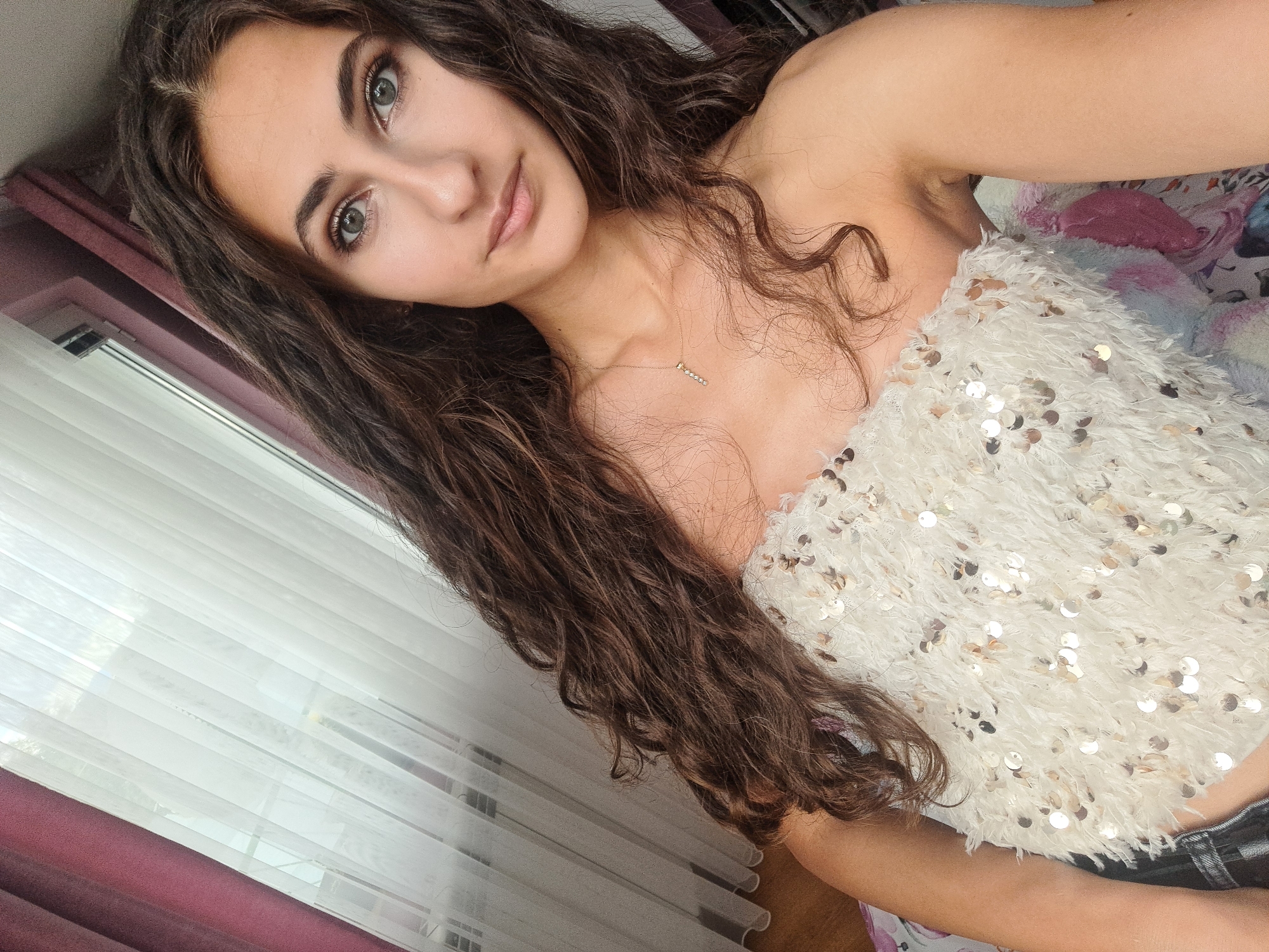 A young woman with long, dark, curly hair is taking a selfie indoors, angled to her left. She is wearing a strapless, white, sequined top. Her left arm is visible, and she is wearing a delicate gold necklace with a small pendant. The background shows a window with sheer white curtains and darker maroon drapes to the left. The lighting suggests daytime. The scene is not identifiable as being in the city of Negovan, Bulgaria.