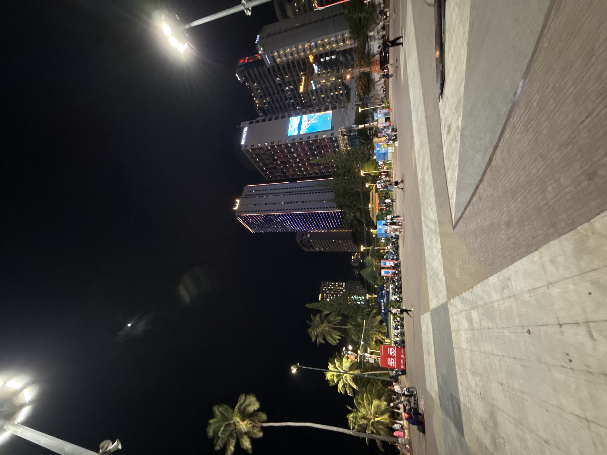 The image depicts a nighttime scene in Nha Trang, Vietnam, likely on the city's popular beach promenade. In the foreground, a wide, tiled pedestrian area is visible, with some sections appearing damp or reflective. A tall lamppost with multiple lights extends into the dark night sky from the left side of the frame, casting a bright illumination. Palm trees, characteristic of the tropical location, are scattered throughout the scene, with one particularly tall, slender palm tree on the left.
In the mid-ground, a bustling area is observed with several modern, illuminated buildings in the background. One prominent building features the "NOVOTEL" logo. Another skyscraper is adorned with vertical lines of purple lights. A large digital billboard on one of the buildings displays a bright, colorful image. A line of parked motorcycles is visible on the road or promenade area, with some people walking and gathered around. Two red booths with white "5G" logos are also present.
The background is dominated by the dark night sky, with a faint moon or light source visible in the upper left quadrant. The overall atmosphere suggests a lively urban environment at night, likely a tourist area with significant pedestrian and vehicular traffic. The presence of palm trees and the general ambiance strongly suggest a coastal city in Vietnam.