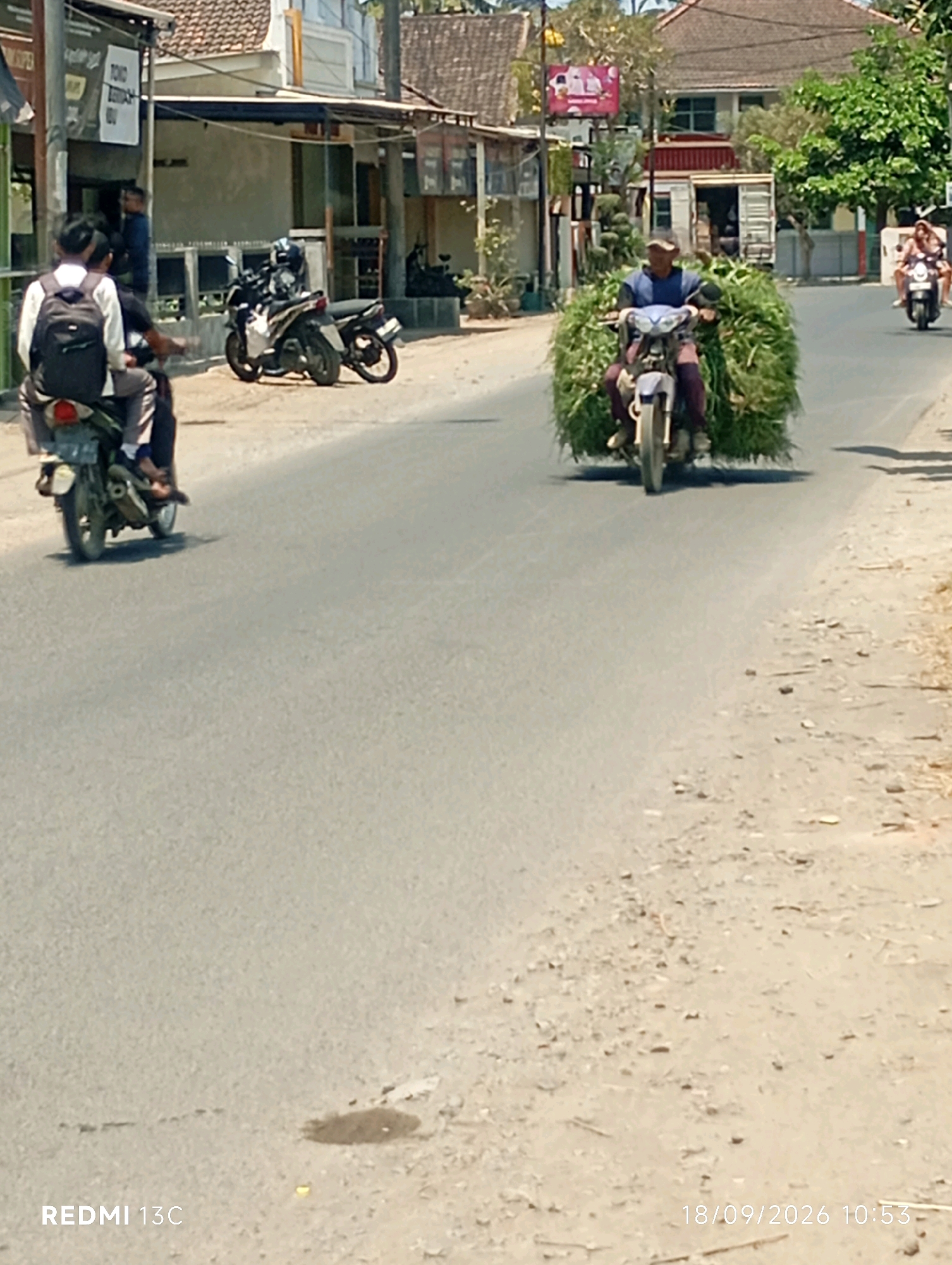 In Sumberrejo, Indonesia, on September 18, 2026, at 10:53 AM, a man rides a motorcycle laden with a large bundle of grass. He is on a paved road, with a dirt shoulder on the right. In the background, there are buildings, trees, and other motorcycles. One motorcycle in the foreground carries two passengers, and another motorcycle is parked in front of a building. The sky is clear and sunny.