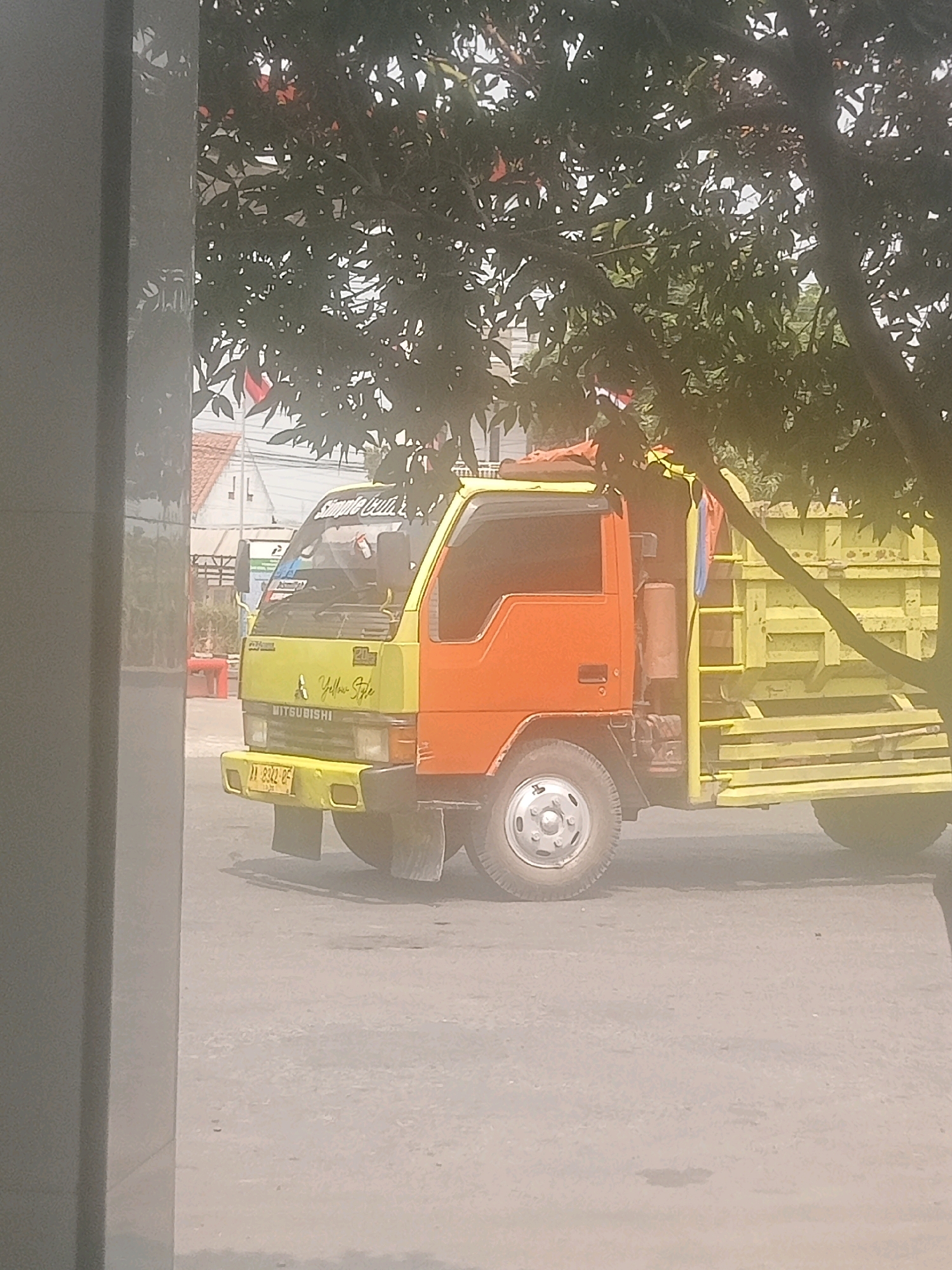 The image shows a two-tone truck, yellow and orange, parked on an asphalt surface. The truck has a yellow bed and a yellow and orange cab, with "Yellow Style" written on the side of the cab in yellow lettering. The truck's license plate is visible and reads "11 2312-CF". In the background, there are buildings and trees, with a flag visible on one of the buildings. The scene appears to be in a city, likely Lebaksiu, Indonesia, given the context. The overall lighting suggests it is daytime, with some haziness in the air.