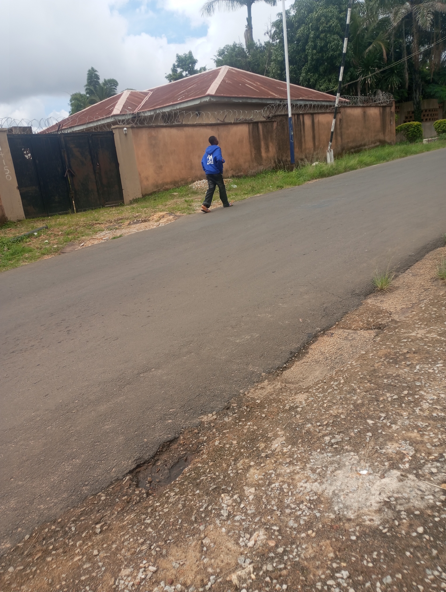 A young person wearing a blue hooded sweatshirt and dark pants walks along the side of a paved road. The person is facing away from the camera and walking towards the right of the image. A low, tan-colored wall with barbed wire along the top runs parallel to the road. Behind the wall are two houses with red roofs and a large dark metal gate. Trees and vegetation are visible behind the houses and wall. The sky is partly cloudy. The road surface is asphalt and shows some signs of wear and tear, with a small pothole visible in the foreground. The area adjacent to the road is covered in grass and gravel. The image appears to have been taken outdoors on a cloudy day. The scene is consistent with a residential area in Jos, Nigeria, likely a suburb or neighborhood due to the presence of a wall and gate surrounding the properties.