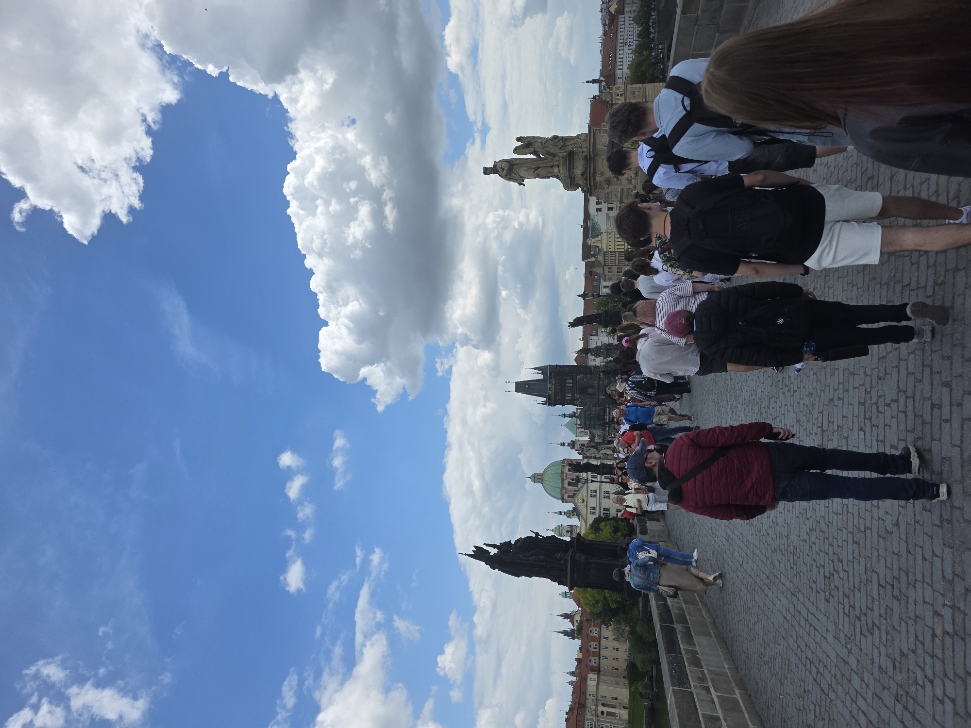The image shows a view from the Charles Bridge in Prague, Czech Republic. The sky is partly cloudy with blue sky visible. A crowd of people are walking on the bridge. Statues line the bridge. Buildings are visible in the background. The bridge is paved with cobblestones.