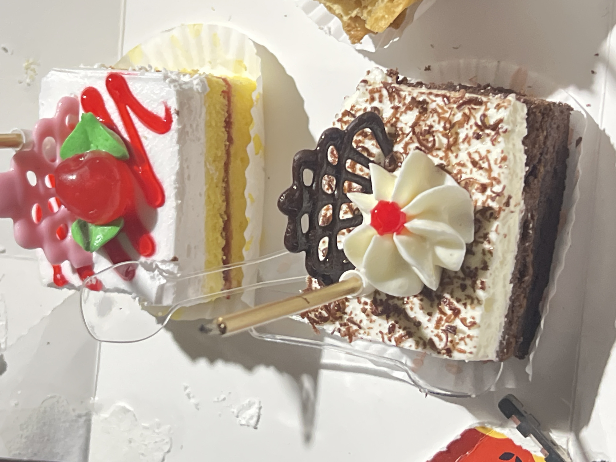 The image displays two slices of cake, positioned in a white box. The foreground features a slice of chocolate cake topped with white frosting, grated chocolate, a decorative white frosting flower, a red cherry, and a single unlit birthday candle. Behind it, to the left, is another slice of cake with white frosting, yellow cake layers, and red and green frosting decorations, including a red cherry. A partially visible pastry is in the upper right corner. The context of Denpasar, Indonesia, is not depicted in the image.