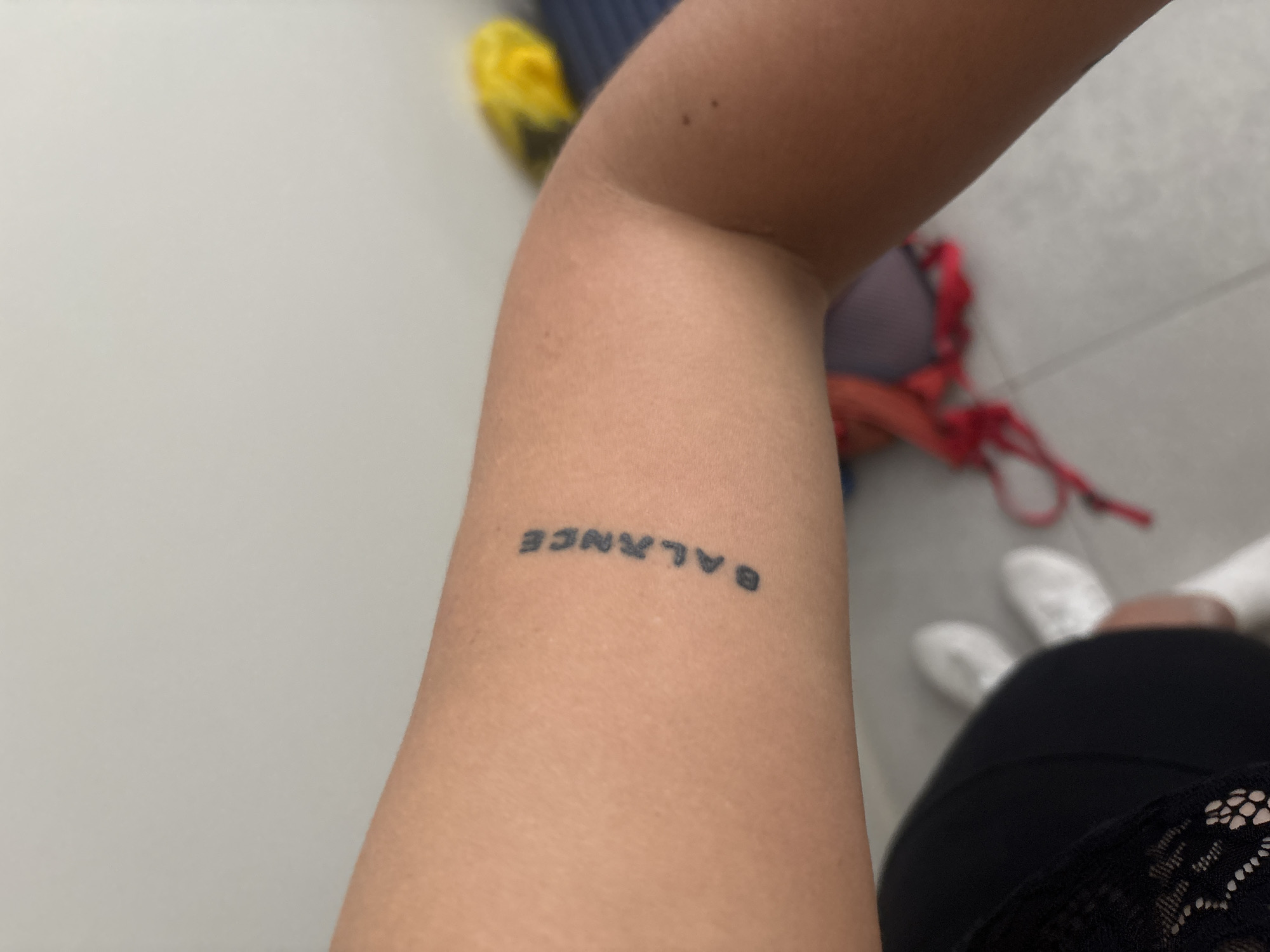 The image displays a close-up of a person's forearm with a tattoo that spells out the word "BALANCE" in a vertical, slightly curved orientation. The tattoo is black and appears to be done in a simple, sans-serif font. The skin tone is light. In the background, out of focus, are some items of clothing, including a black top with lace detailing, white sneakers, and a bright yellow object, possibly a bag or piece of clothing. There are also hints of a blue surface. The setting appears to be indoors, possibly a locker room or a changing area, given the presence of footwear and athletic-style clothing. There is no indication of the city of Ldzaa, Abkhazia in the image.