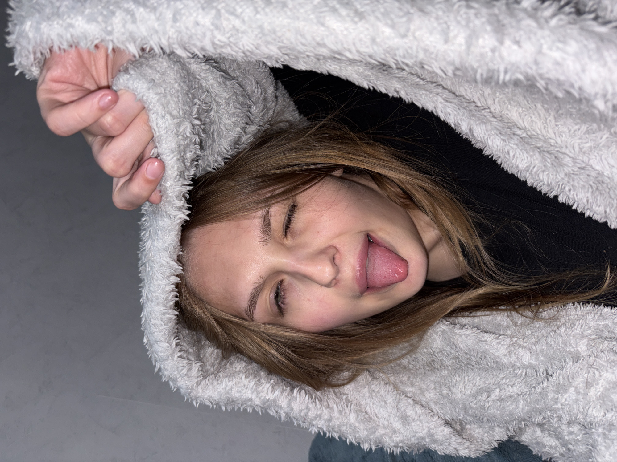 An angled, close-up portrait features a young individual, likely female, captured in a playful moment. Her head is tilted, with eyes mostly closed or squinting, and her tongue is sticking out in a whimsical gesture, suggesting a lighthearted and silly mood. She has straight, light brown hair that falls around her face.
The person is partially enveloped by a thick, fuzzy, light-colored (appearing white or very light grey) hooded garment or blanket, which dominates a significant portion of the frame. A dark, possibly black, garment is visible underneath the fuzzy material. One hand, with neatly manicured nails featuring a natural or clear polish, is seen holding onto the textured fabric at the upper left. The background is a plain, light grey surface, indicating an indoor setting with even, diffused lighting. No text is visible. The scene evokes a sense of casual comfort and youthful cheerfulness. This image is contextualized as being from Chaikovsky, Russia.