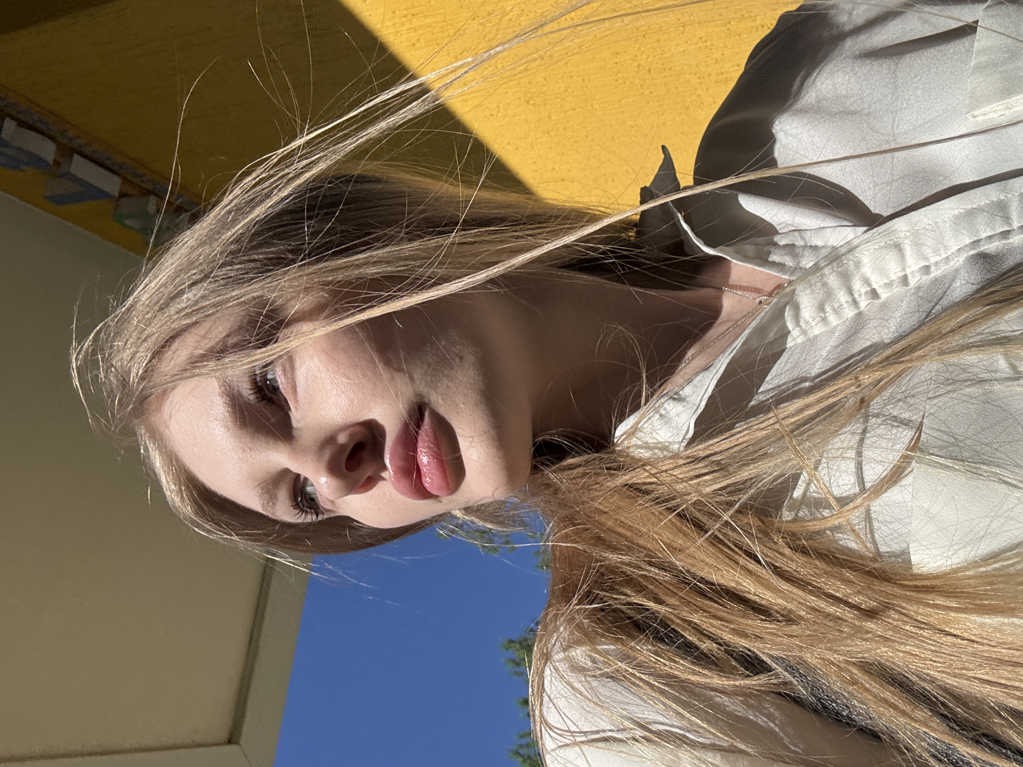 A low-angle photograph shows a young woman with long, blonde hair posing in what appears to be an outdoor location. The background features a yellow-painted surface or wall with a shadow cast across it, along with a glimpse of blue sky and green foliage. The woman is wearing a light-colored jacket or shirt. The lighting suggests it is daytime, with natural light illuminating the scene. The location is identified as Perm, Russia, based on the information provided.