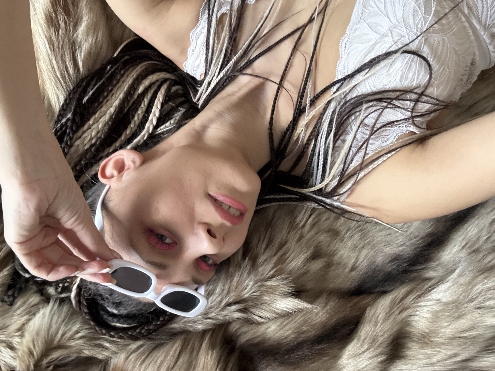 A woman with braided hair, wearing a white lace top, is lying on a furry surface. She is holding a pair of white-framed sunglasses near her face. The scene appears to be indoors. The location information, Rusanovo, Russia, is extraneous to the observable content of the image.