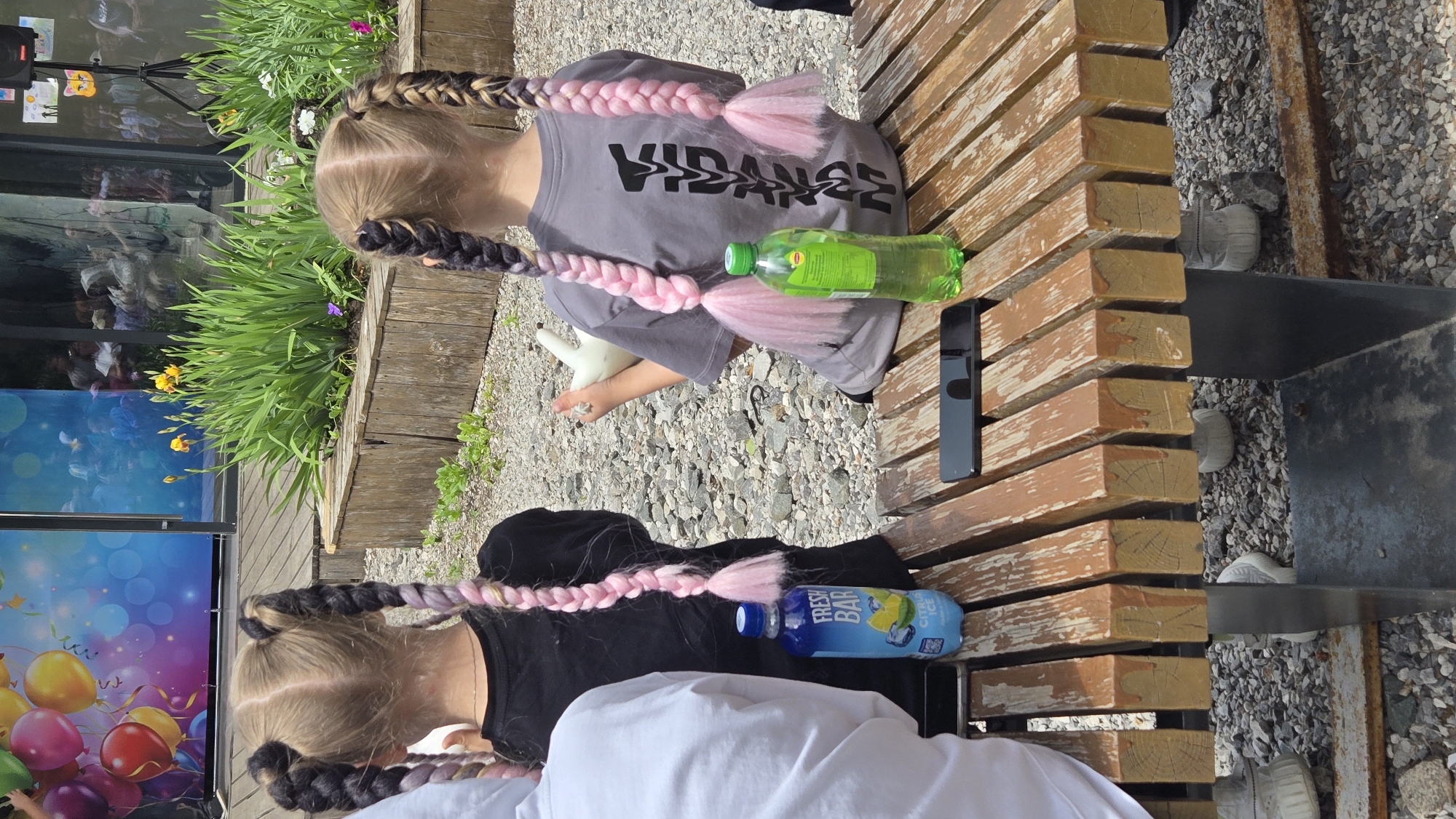 Two young individuals are positioned on a wooden bench, with a gravel surface visible beneath. The first individual is wearing a grey t-shirt with the text "VIDANGE" printed on it and has their hair styled in braids adorned with pink extensions. A green water bottle rests next to them on the bench. The second individual is clad in a black top, partially obscured by a white shirt or jacket. They also sport braids with pink extensions. A blue beverage bottle is situated beside them. A black mobile phone is placed between the two individuals on the bench. In the background, greenery, possibly a garden or park area, is visible, along with a wall or structure with a colorful display or decoration. The location appears to be an outdoor public space, possibly a park or recreational area.