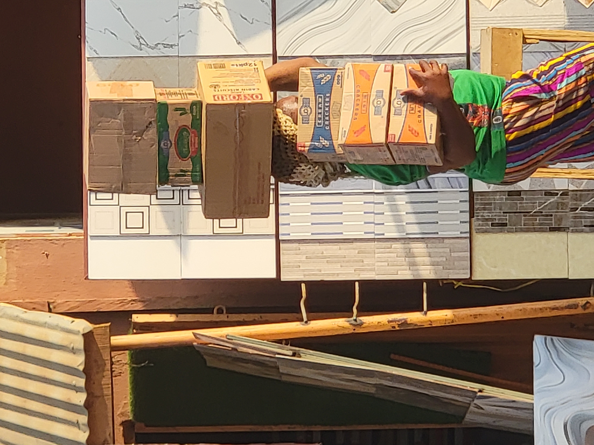 In Maiburiji, Nigeria, a person is holding boxes of "Cream Crackers" and "Oxford Cabin Biscuits" in front of a tile display. The person is wearing a green shirt and a colorful striped garment. The tiles are various patterns and colors, arranged in a grid. The scene appears to be outdoors, with wooden structures visible in the foreground.