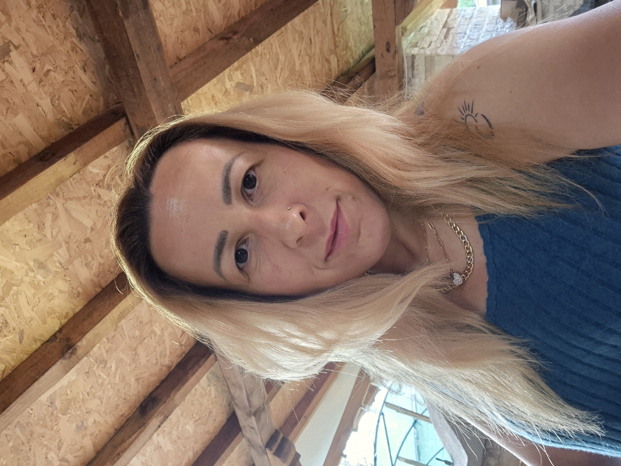 A woman with blonde hair is taking a selfie. She is wearing a blue top and gold necklaces. She has a tattoo of a sun and moon on her shoulder. The background is a wooden ceiling with exposed beams.