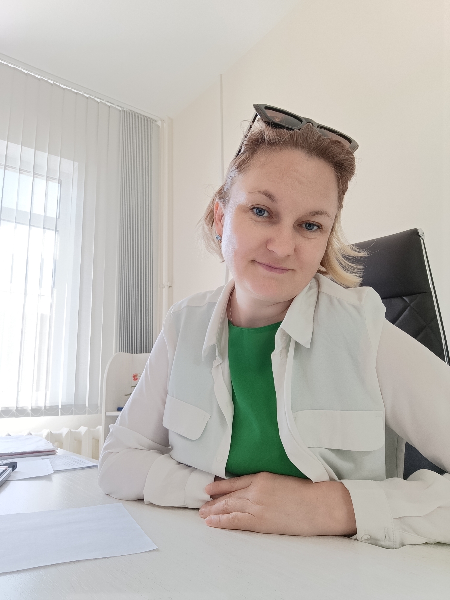 A woman is seated at a desk in an office in Марковский, Russia. She has blonde hair and is wearing a white shirt over a green top. Sunglasses are perched on her head. The desk is made of white wood and has papers and a calculator on it. Behind her is a black office chair, and to her left is a window with vertical blinds. The room appears to be well-lit by natural light.