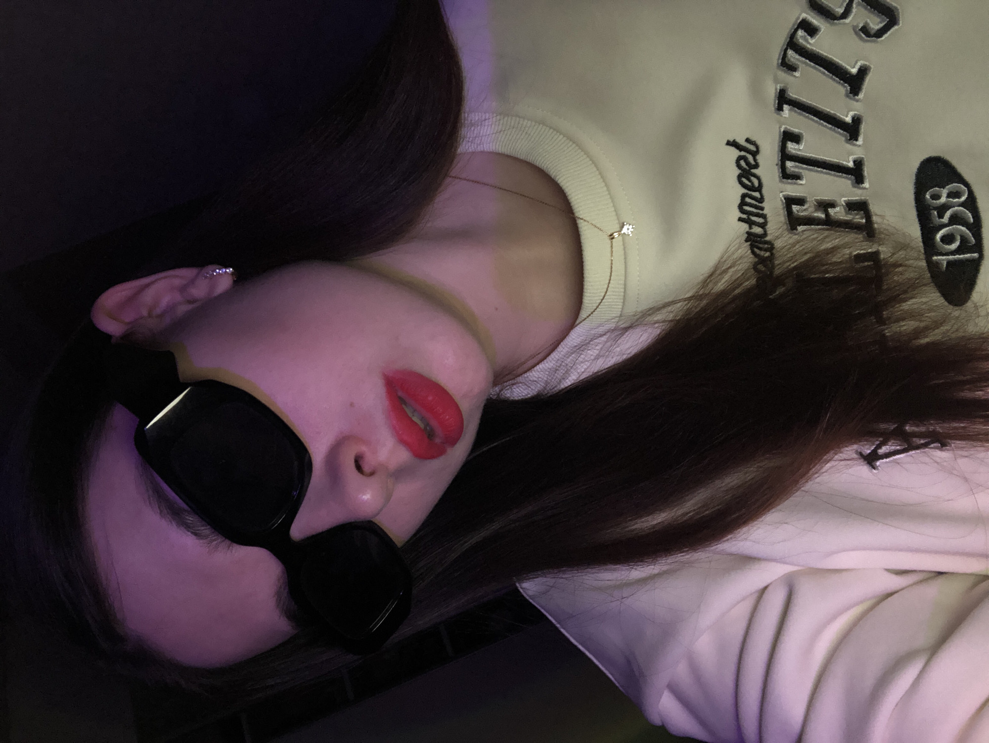 A young woman is depicted in a close-up, horizontally oriented shot, appearing to lie down indoors in Noviy, Russia. Her long, dark brown hair frames her face and spreads across a light-colored sweatshirt.
She wears black, square-framed sunglasses that cover her eyes, and striking bright red lipstick on her slightly parted lips. A delicate gold chain necklace with a small, sparkling pendant rests on her neck, partially visible above the ribbed collar of her sweatshirt. On her visible ear, a small, shiny helix piercing can be seen.
The sweatshirt, possibly cream or pale yellow, features embroidered text. Visible words include a cursive text, likely "heartmeet" or "apartment," followed by large block letters "FITT" (with subsequent letters partially obscured). Below this, the year "1958" is clearly displayed within a dark oval patch.
The setting appears to be a dimly lit room, illuminated by artificial light that casts a soft purple hue on her skin and the surrounding background, creating a moody atmosphere. The overall impression is one of a relaxed, stylish pose.