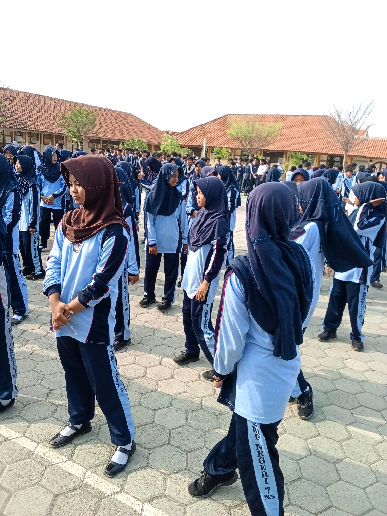 A group of young students, predominantly female, are gathered outdoors in what appears to be a schoolyard. They are wearing school uniforms consisting of light blue long-sleeved shirts with navy blue accents and navy blue pants. Many of the female students are also wearing navy blue hijabs. The ground is paved with hexagonal interlocking tiles. In the background, there are buildings with red tile roofs, suggesting an Indonesian setting, possibly a school campus. The students are facing various directions, some standing still, others in motion, implying they might be participating in an activity or assembly. The lighting indicates it is daytime, with a bright sky suggesting clear weather. One student in the foreground, wearing a brown hijab and a light blue uniform, is looking to her left. The overall scene depicts a typical school environment in Indonesia.