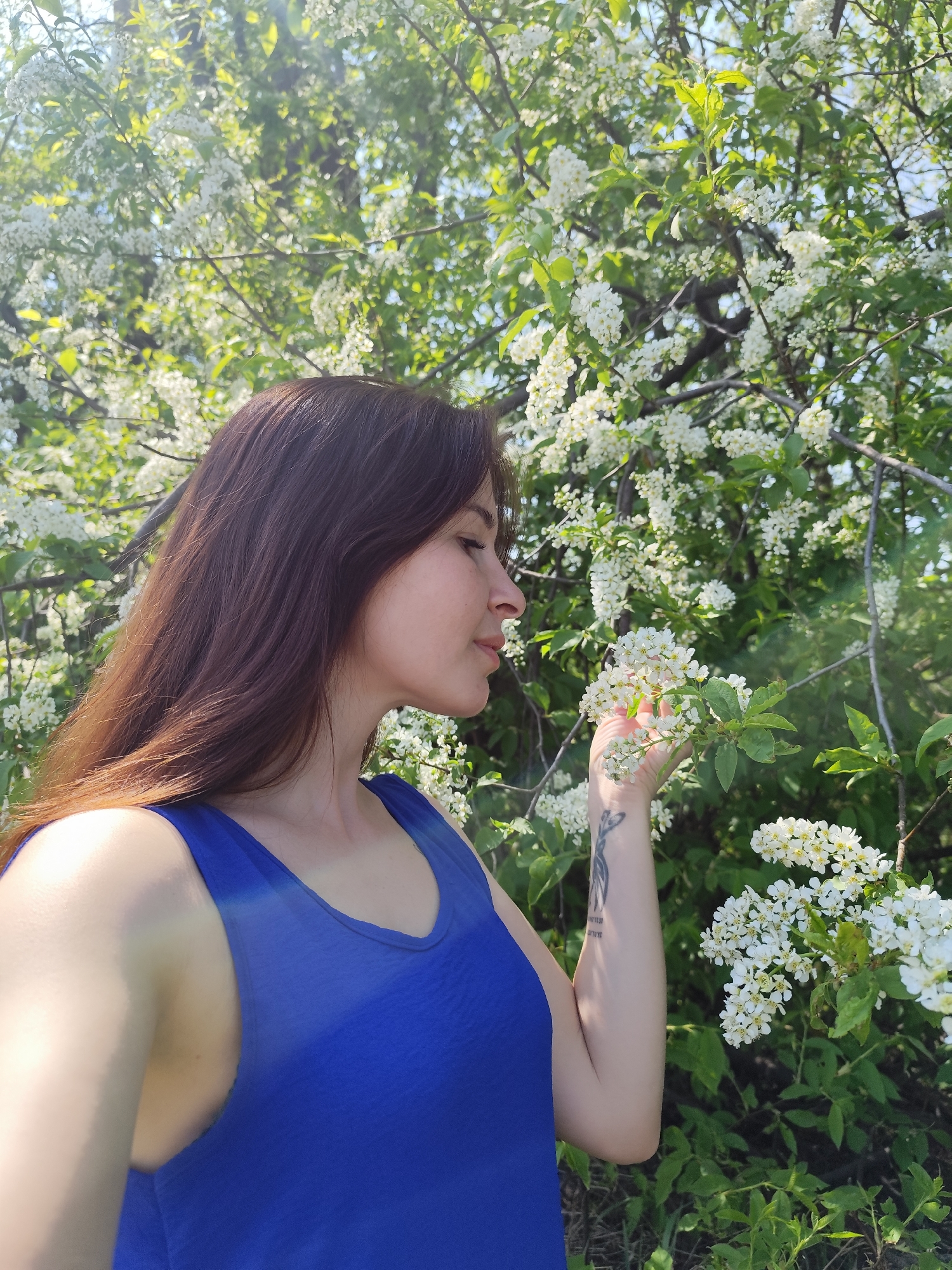 A woman in a blue tank top is standing in front of a flowering tree, possibly in a park or garden setting. The woman is looking at the flowers, holding a branch in her hand. She has long brown hair and a tattoo on her arm. The background is filled with white flowers and green leaves. The location is Фоки, Russia, based on the prompt.