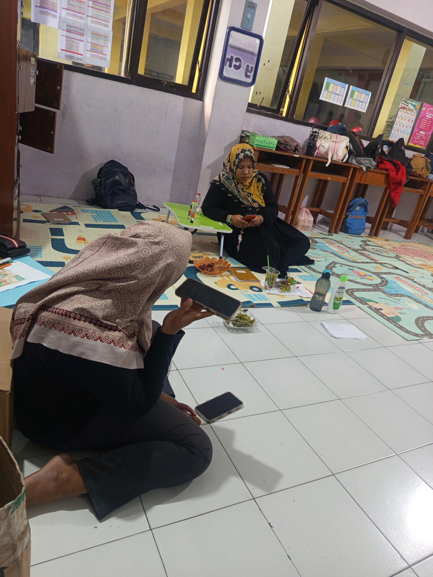Two women are sitting on the floor in a classroom or common area. The woman on the left is mostly obscured, facing away from the camera, holding a mobile phone. She is wearing a headscarf and dark clothing. The woman on the right is wearing a black abaya and a yellow and black patterned headscarf, looking down at her mobile phone. She is sitting on a mat with a colorful children's cartoon theme. In front of her is a small green table with snacks and drinks. There are wooden tables and chairs in the background, with windows showing a bright exterior. A backpack is on the floor near the wall. The overall setting appears to be an educational or community space, possibly a madrasah or school in Malang, Indonesia.