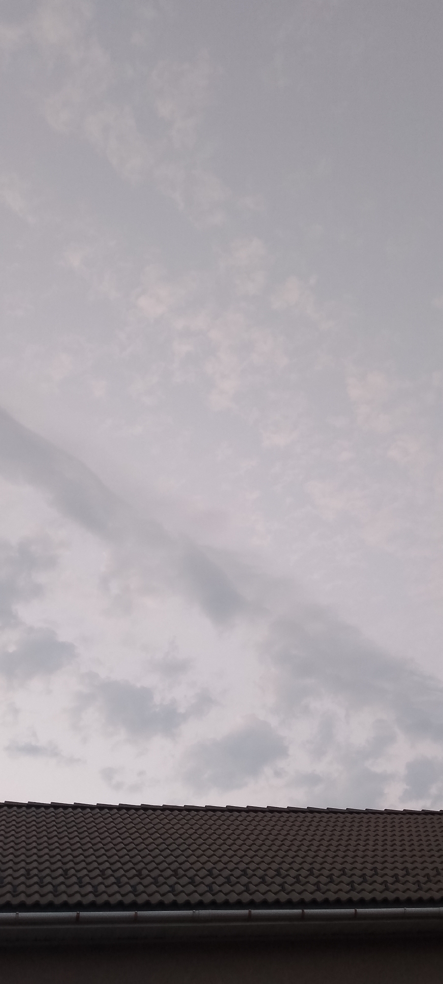 The image shows a close-up, eye-level, vertical shot of a gray, cloudy sky above a dark-tiled roofline. The roof tiles are arranged in a repeating pattern of curved shapes. A silver gutter runs along the bottom edge of the roof. The sky is filled with various shades of gray clouds, with some lighter areas indicating the presence of sunlight attempting to break through. The overall impression is of an overcast day in an urban environment, possibly a residential area. No people or specific landmarks of Belgorod, Russia, are visible in this cropped image.