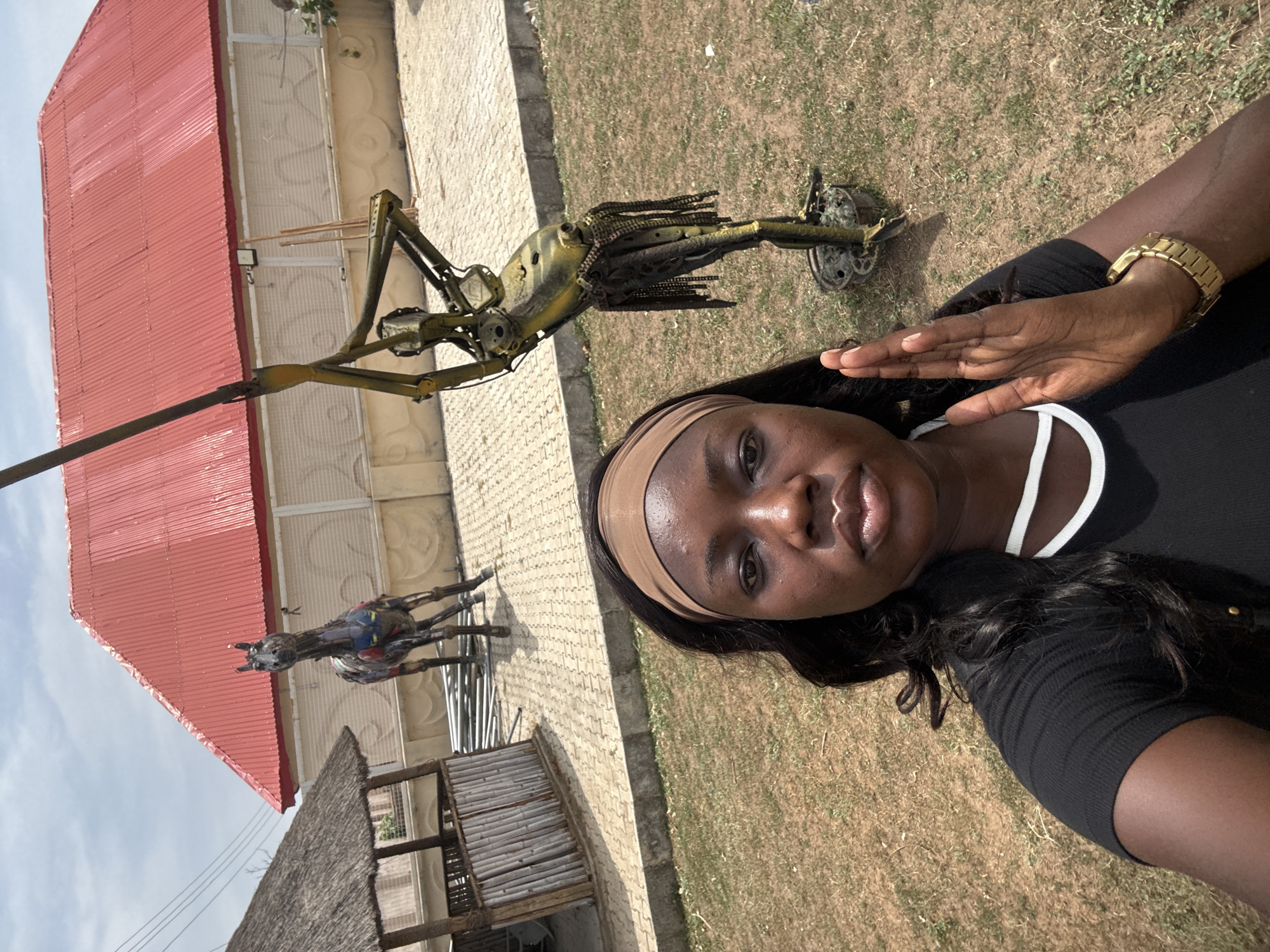 A woman is featured in a selfie taken outdoors in Abuja, Nigeria, with artistic sculptures and architectural elements in the background. She has dark skin, long dark hair, and wears a light brown headband, a black ribbed top, and a gold-colored wristwatch on her left wrist. Her expression is neutral, and her left hand is gently raised.
Behind her, a prominent, tall metallic sculpture of a human-like figure stands on a small wheeled base. This sculpture appears to be constructed from various recycled industrial components such as chains and gears, exhibiting a yellowish-green metallic finish. Further in the background, a second, multi-colored metallic sculpture resembling a horse is visible.
The setting includes a building with a striking red corrugated metal roof, occupying a significant portion of the background. Another smaller structure with a thatched roof is partially visible lower in the background. The ground is a mix of dry grass and paved areas with interlocking stones. The bright, clear sky and strong natural light indicate that the photo was taken during the daytime. No visible text is present in the image.
