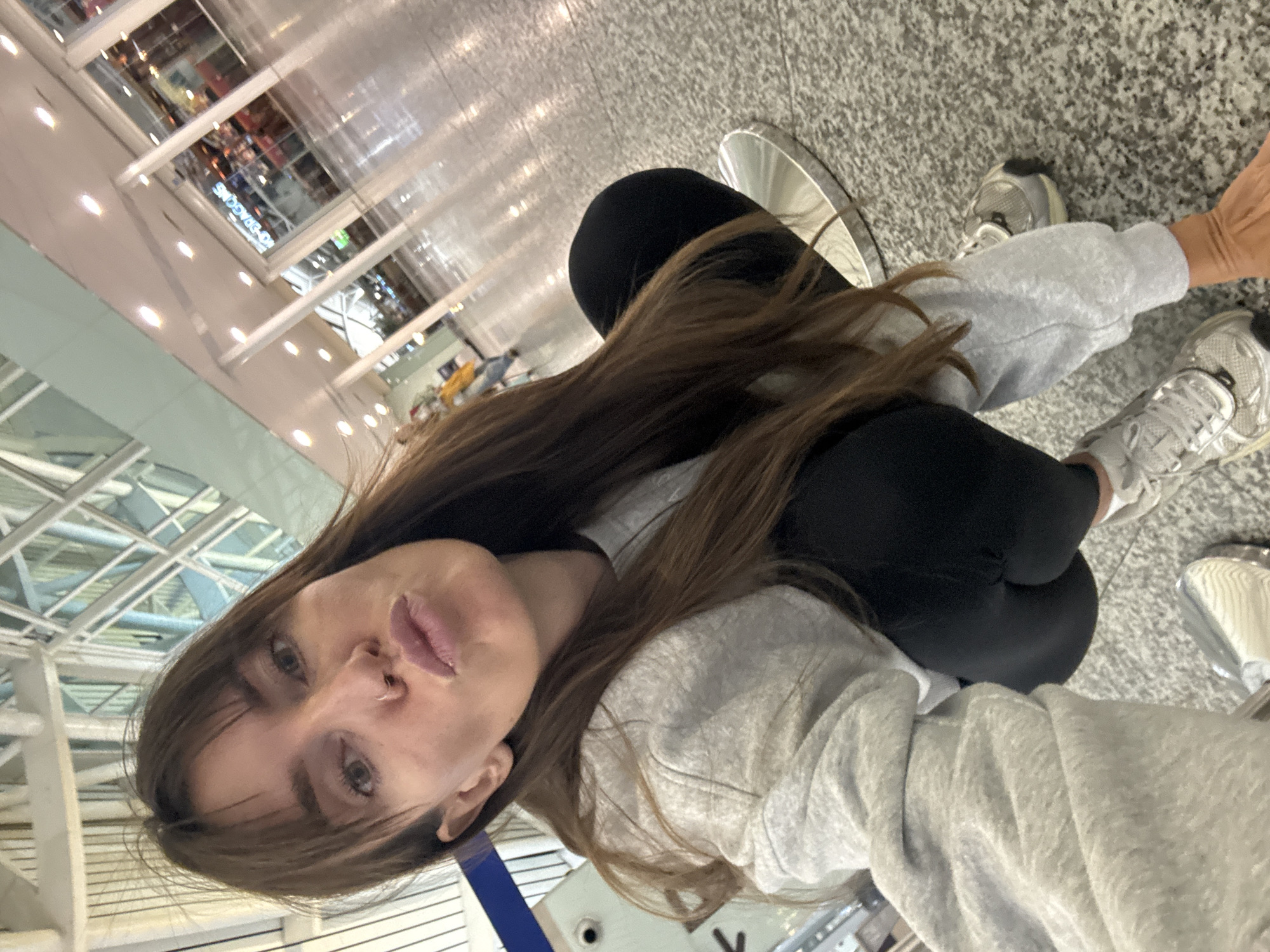 A woman with long brown hair and bangs, wearing a grey hoodie and black leggings, crouches on a speckled grey floor. She is looking at the camera with her lips pursed. Her right hand is extended, resting on the floor. She is wearing silver sneakers. In the background, there is a modern building with large windows and bright lights. The environment appears to be an indoor public space, possibly an airport terminal or a shopping mall, with a polished floor reflecting the overhead lighting. The image does not provide any specific information that would place it in Kuta, Indonesia.