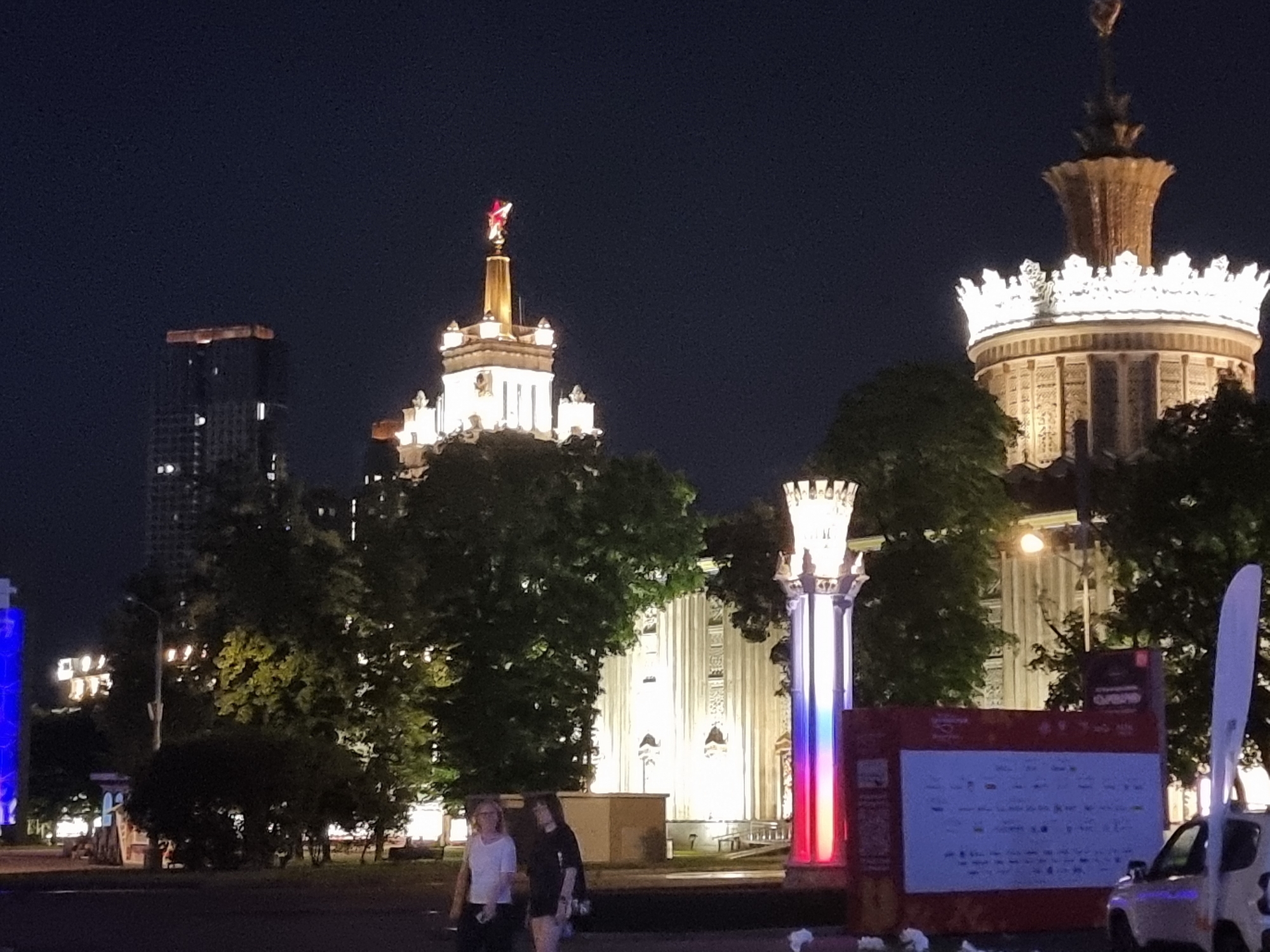 This is a nighttime photograph of the All-Russian Exhibition Center (VDNKh) in Moscow, Russia. The image features several iconic pavilions illuminated against the dark sky. On the left, a modern skyscraper with illuminated windows stands tall. In the center, the main pavilion of VDNKh, topped with a red star, is brightly lit. To the right, another grand pavilion, with ornate decorations and a crown-like spire, is also illuminated. Trees are scattered throughout the scene, some obscuring parts of the pavilions. In the foreground, a street lamp is visible, and two women are walking away from the camera. A modern, colorful illuminated column stands to the right of the women, and a large red and white banner with text and logos is also present. A white vehicle is parked to the right of the banner. The overall atmosphere is festive, likely due to the nighttime illumination of the exhibition center.