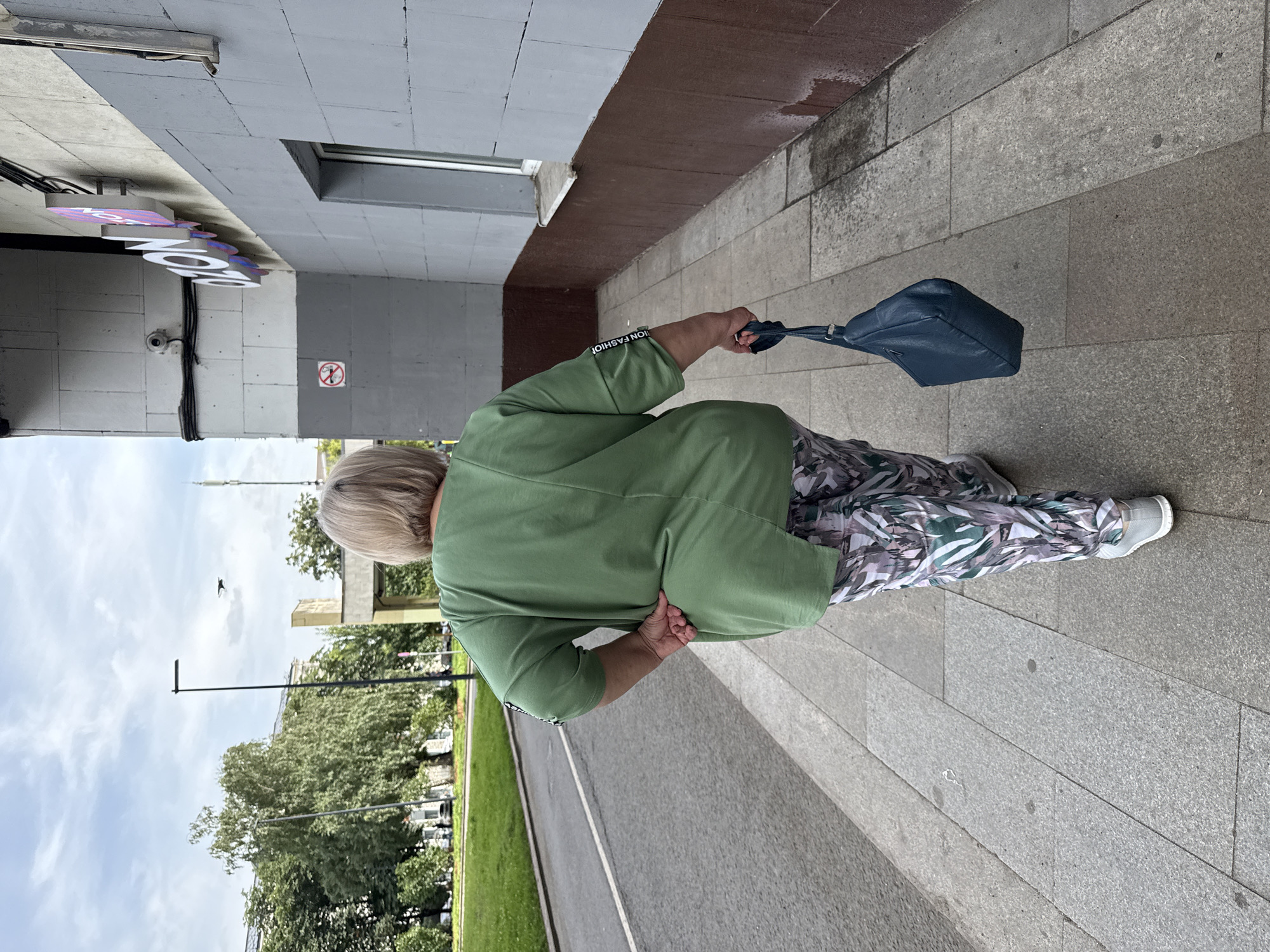 A full-shot, rear view of a Caucasian woman walking on a sidewalk in Moscow, Russia. The woman is wearing a green, oversized tunic top and patterned pants. She is holding a dark blue purse in her right hand. The sidewalk is made of concrete and is bordered by a street on the left and a building on the right. The building has a grey facade with a brown base and a window. A sign that says "OZON" is visible on the building. The sky is cloudy.