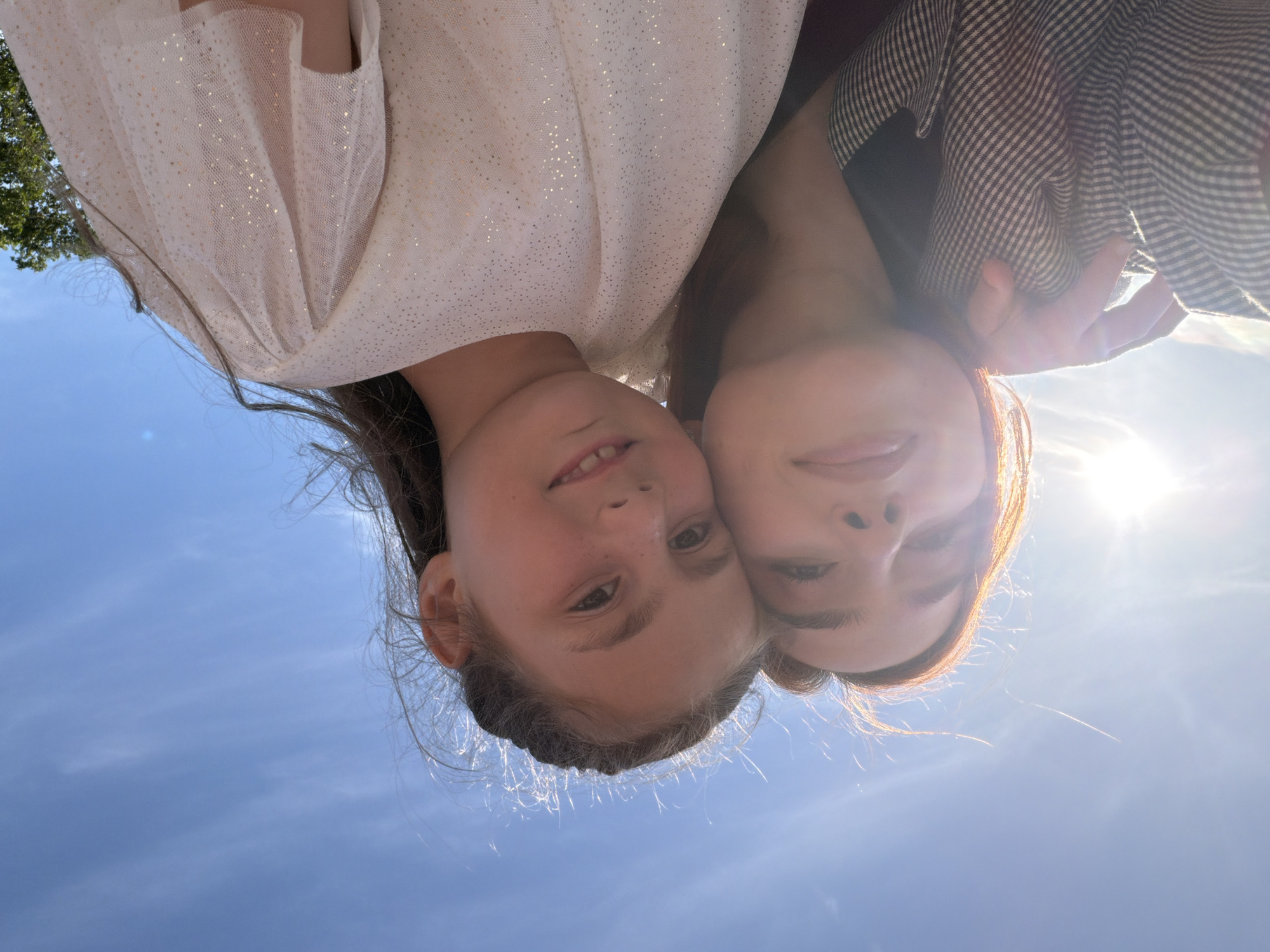 Two individuals, one appearing to be a young girl and the other an older female, are captured in a close-up selfie outdoors. The sky is a clear, bright blue with wispy clouds and the sun is visible in the upper left corner, creating a lens flare effect. The older female, on the left, has reddish-brown hair and is wearing a plaid jacket. She has her arm around the younger girl. The younger girl, on the right, has dark hair styled in braids and is wearing a white top with gold speckles. She is smiling slightly. The image composition is tight, focusing on their faces and upper bodies, with the sky dominating the background. The scene does not provide any specific visual cues to indicate it is located in Strasbourg, France.
