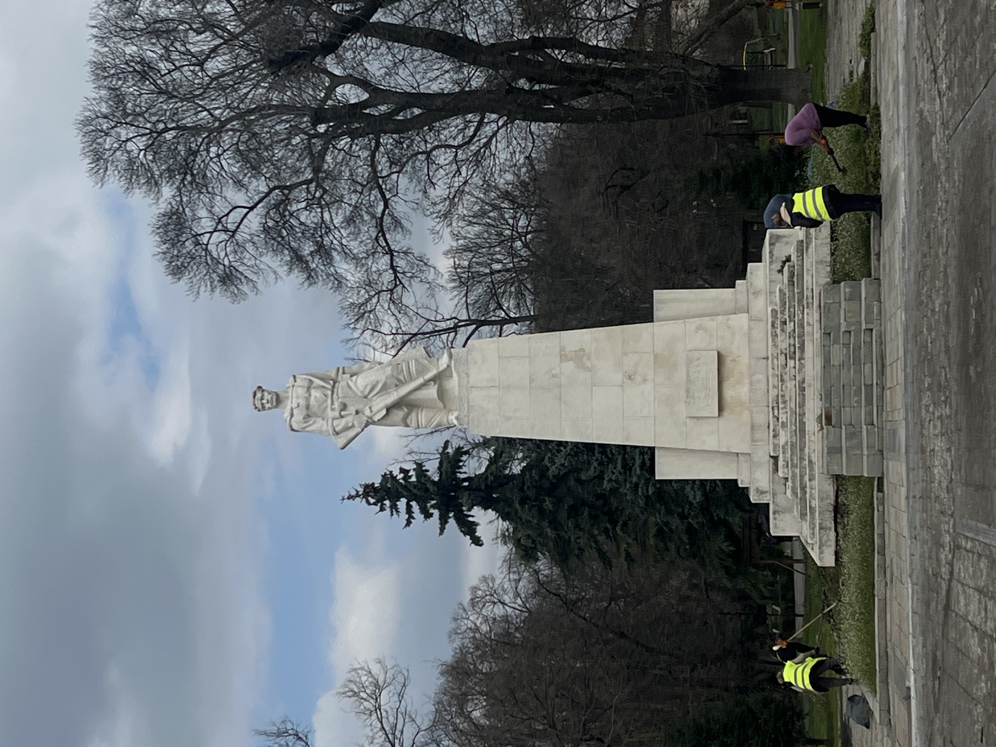 The image depicts a white stone monument in Karnobat, Bulgaria. The monument features a sculpted soldier holding a rifle. Two people wearing yellow vests are tending to the landscaping at the base of the monument. A third person in a pink top is also visible in the background. The monument is situated in a park-like setting, with trees and cloudy sky visible. The image appears to have been taken during the day.