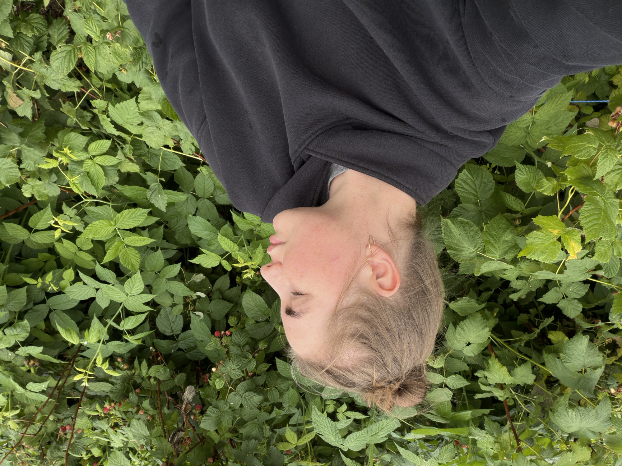 An individual with blonde hair in a bun is shown from the chest up, in profile facing right. They are wearing a dark grey hooded sweatshirt. The person is situated in front of a dense green bush with visible red berries. The background is primarily foliage, suggesting an outdoor setting, possibly a garden or park in Уральское, Russia. The lighting appears natural and diffused, consistent with an overcast day.