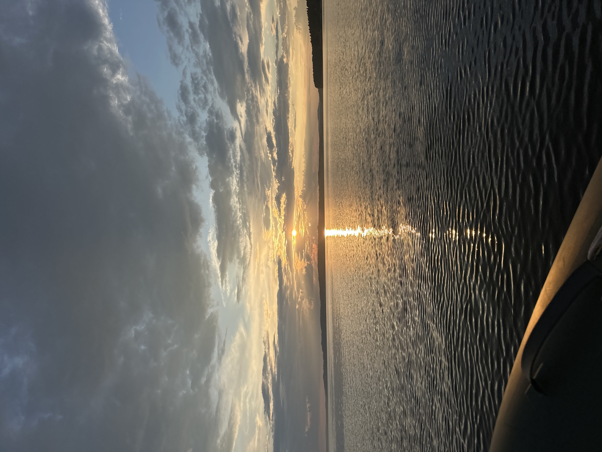 The image captures a serene sunset over a body of water, likely a lake or river, near Sosnovka, Russia. The sky is a dramatic canvas of gray and white clouds, with the setting sun casting a warm, golden glow in the center, creating a shimmering reflection on the water's surface. The horizon is marked by a dark, tree-lined shore. In the foreground, the edge of a dark inflatable boat is visible, suggesting the photograph was taken from its perspective. The water is calm, with small ripples distorting the reflection of the sun. The overall atmosphere is tranquil and picturesque, indicative of a peaceful evening by the water.
