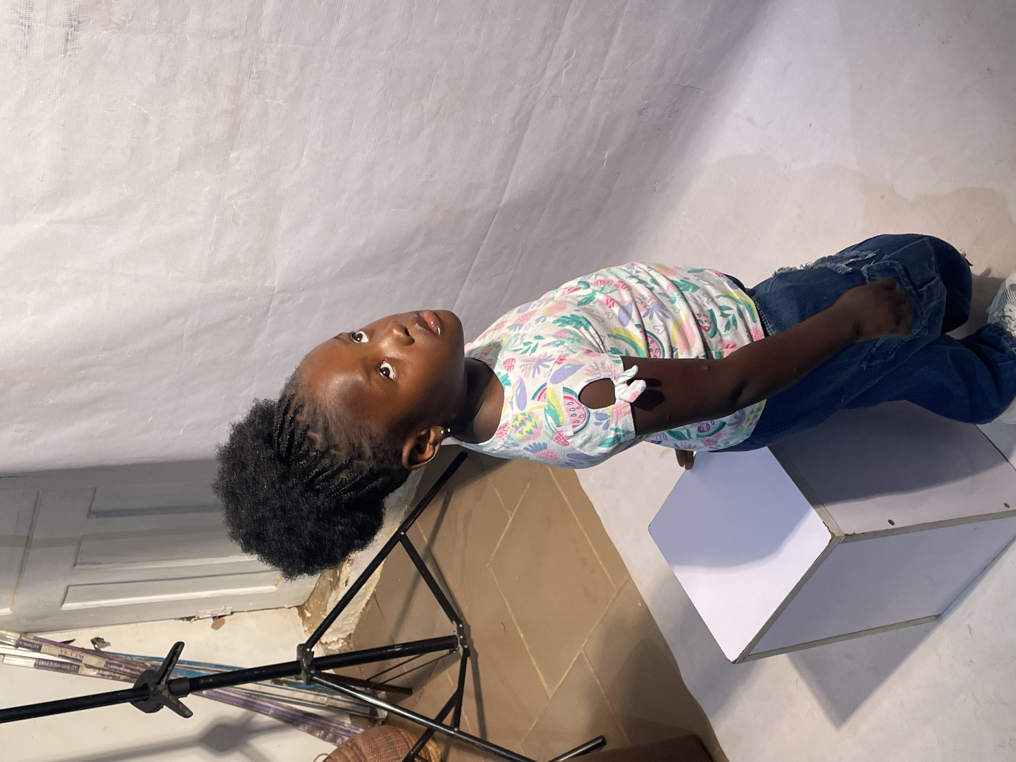 A young Black girl with braided hair styled in a high puff is posing for a photograph. She is wearing a white t-shirt with a colorful fruit pattern and blue jeans. She is standing next to a white cube, and behind her is a light-colored backdrop. To her left, a photography light stand is visible. The environment appears to be an indoor studio setting. The image was captured in Jos, Nigeria.