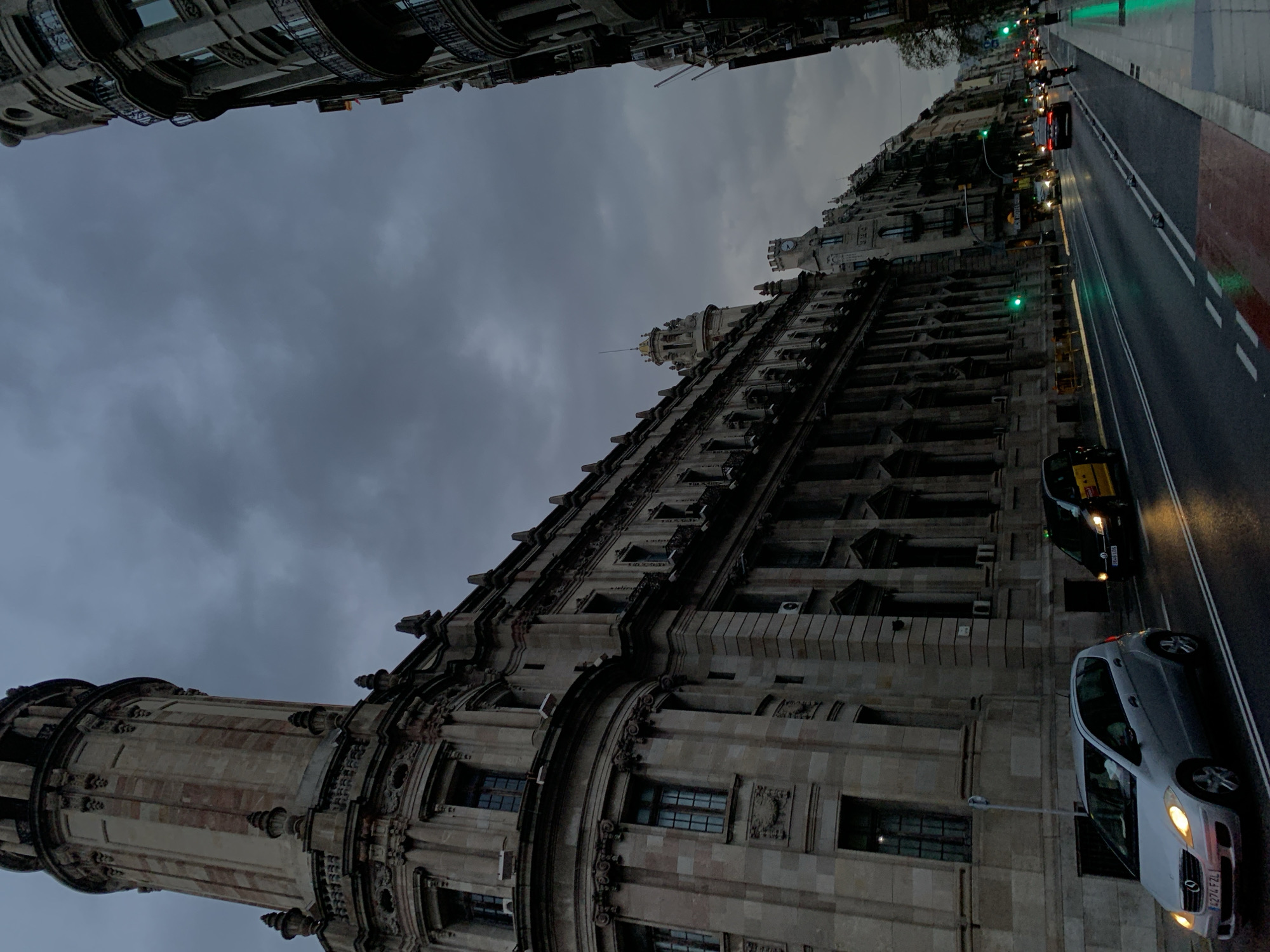 The image captures a street view in Barcelona, Spain. Dominating the frame is the façade of a historic building, likely a government or public building, with ornate stone architecture, including a tower and many windows. The building's lower levels are visible, with a tower featuring multiple sections. The cloudy sky overhead casts a muted light. A street runs alongside the building. Vehicles, including a silver car and a yellow taxi, are driving on the road. The street has marked lanes and pedestrian areas. Traffic lights are visible, and indicate it is night, or a low light situation. Buildings across the street mirror similar architectural styles and height. The overall atmosphere is urban and historical.