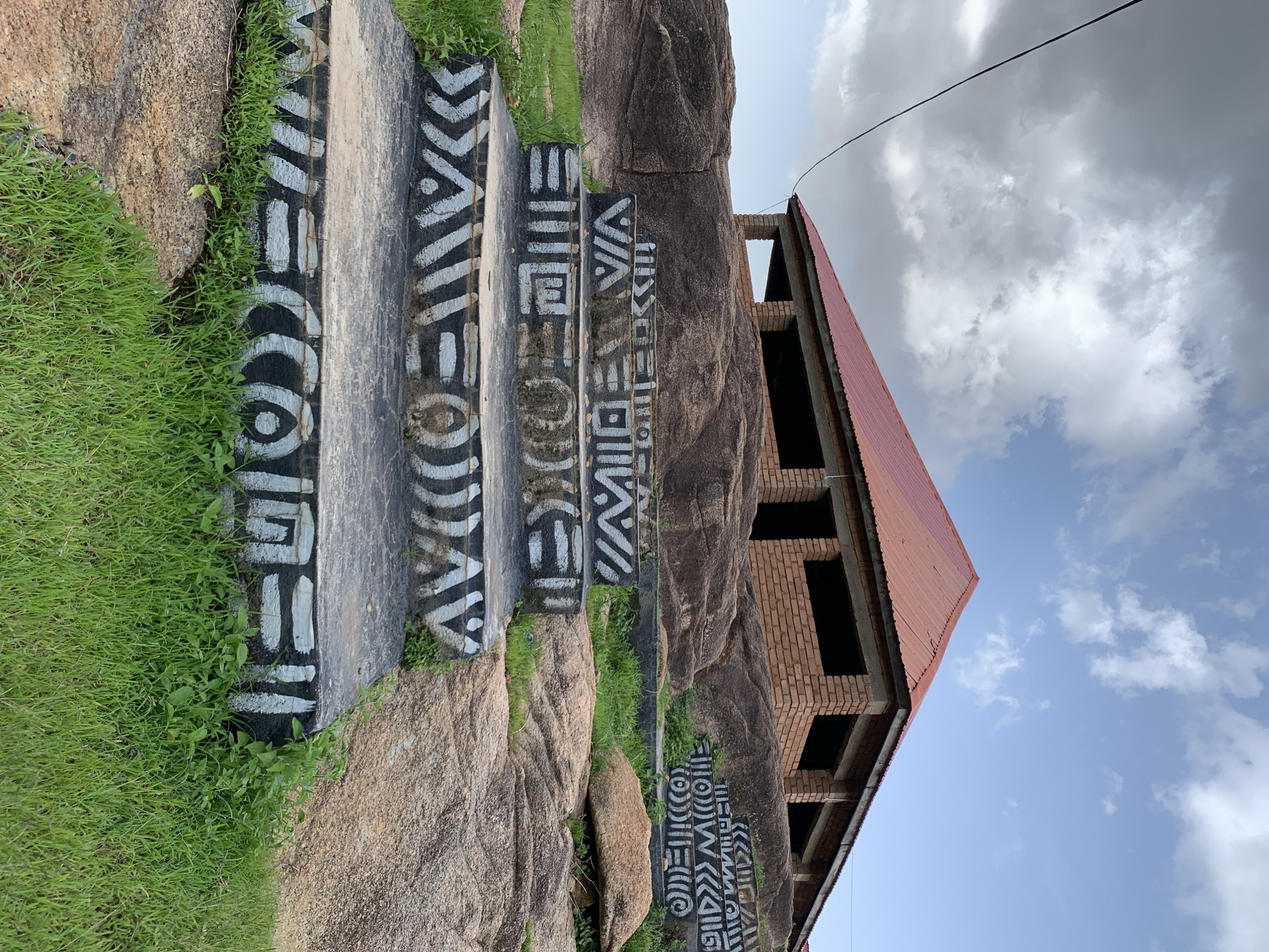 In Jos, Nigeria, a set of concrete stairs, adorned with black and white tribal markings, ascends a rocky incline. The stairs lead towards a partially constructed building with a red corrugated metal roof, its brick walls unfinished and window openings exposed. The surrounding environment consists of large, weathered rocks and patches of green grass. The sky above is a mix of gray clouds and blue patches, suggesting a partly cloudy day. A dark wire, possibly a power line, stretches across the upper left corner of the frame. The scene appears to be an outdoor location, likely a public space or a developing site.