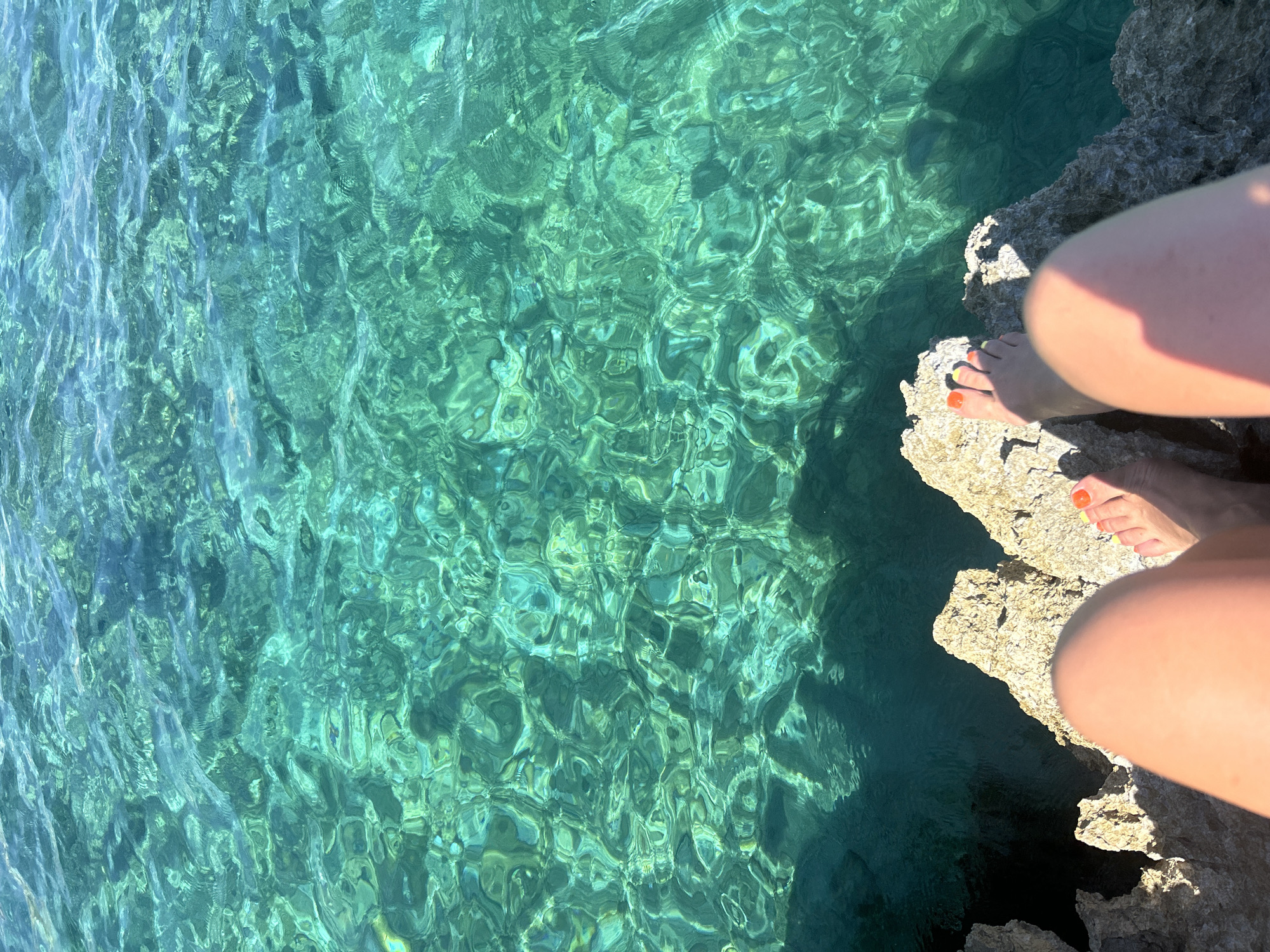 In Cala Lliteres, Spain, a person's bare legs and feet are visible, positioned on a rocky outcrop overlooking the turquoise Mediterranean Sea. The individual's toenails are painted with bright orange and yellow polish. The sunlit water exhibits a ripple effect, creating a mesmerizing pattern of light and shadow on the seabed below. The rocks are textured and appear weathered, characteristic of a coastal environment.