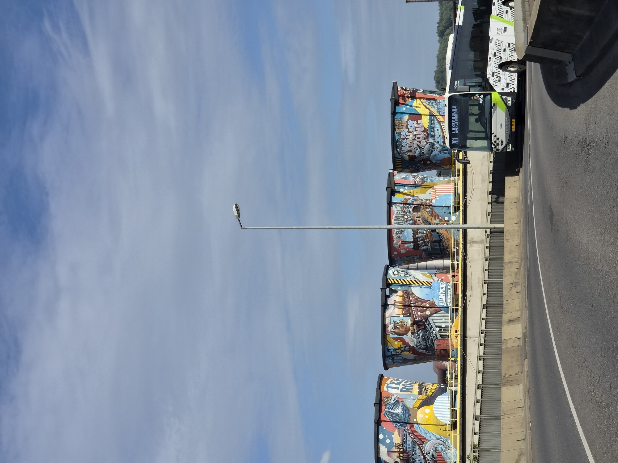 The image depicts the cooling towers in Differdange, Luxembourg, adorned with colorful murals. A white and green bus with the number 701 and "LUXEMBOURG AVENUE MONTEREY" displayed on its front is traveling on a road in the foreground. The bus has "RGTR" visible on its side and a distinctive black and white patterned design. The sky above is partly cloudy with a bright blue hue. The cooling towers, featuring intricate artwork depicting industrial scenes and characters, are prominent in the background, rising above a yellow railing and a grey fence. A street lamp is positioned between the first and second cooling tower from the left. The road in the foreground is asphalt with a white dividing line.