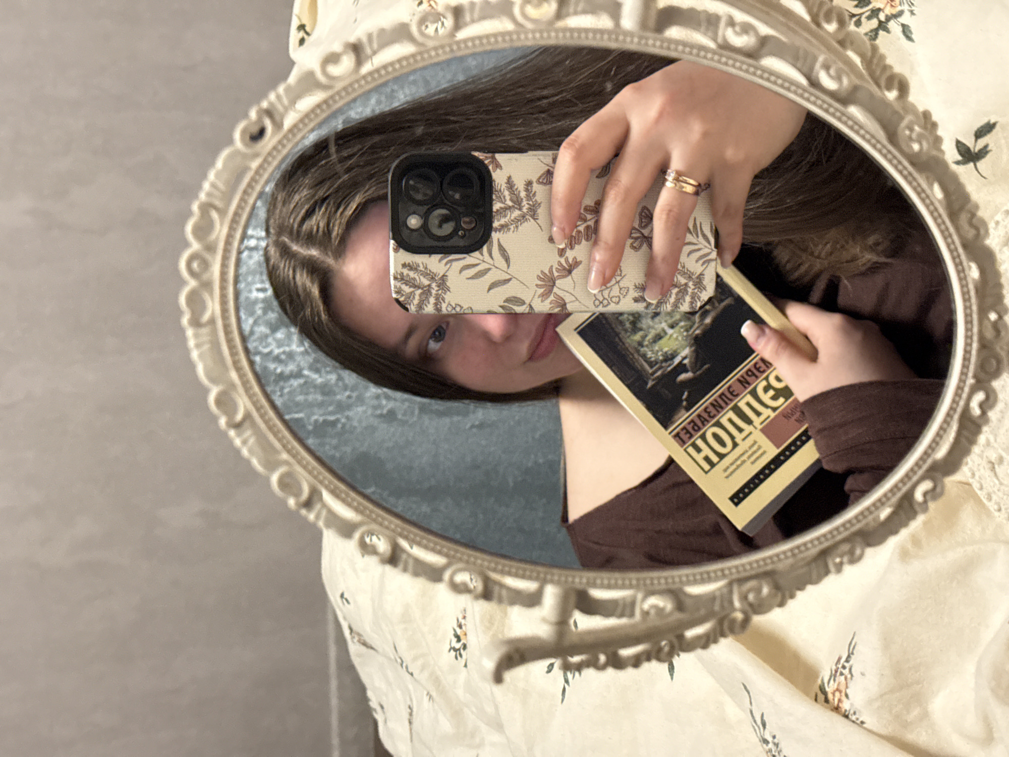 An indoor scene, likely a residential bedroom, presumed to be within Krasnodar, Russia. The image is a mirror selfie captured by a female individual with long brown hair, visible from the chest up. She is wearing a dark brown long-sleeved top. Her left eye is partially visible, directed slightly to her left.
She holds a dark-colored smartphone with a light-toned, floral-patterned case in her right hand, positioned to capture her reflection in an ornate, off-white framed mirror. The smartphone features a visible triple-lens camera array. On her right ring finger, she wears two gold-colored rings, one with a small clear stone. Her fingernails have light-colored polish.
In her left hand, held upside down and reversed in the mirror, is a book with a yellow/gold cover. The visible Cyrillic title reads "НОЛЛЕС" (Nolles). The book cover also displays a dark illustration, possibly depicting a landscape or building.
Reflected in the mirror behind the individual is a textured light blue or teal surface, possibly a wall covering or curtain. Outside the mirror's frame, to the left and bottom, a light-colored duvet or blanket with a sparse floral pattern of green stems and small orange/brown motifs is visible. Above and to the left of the mirror, a plain, light grey wall is visible.