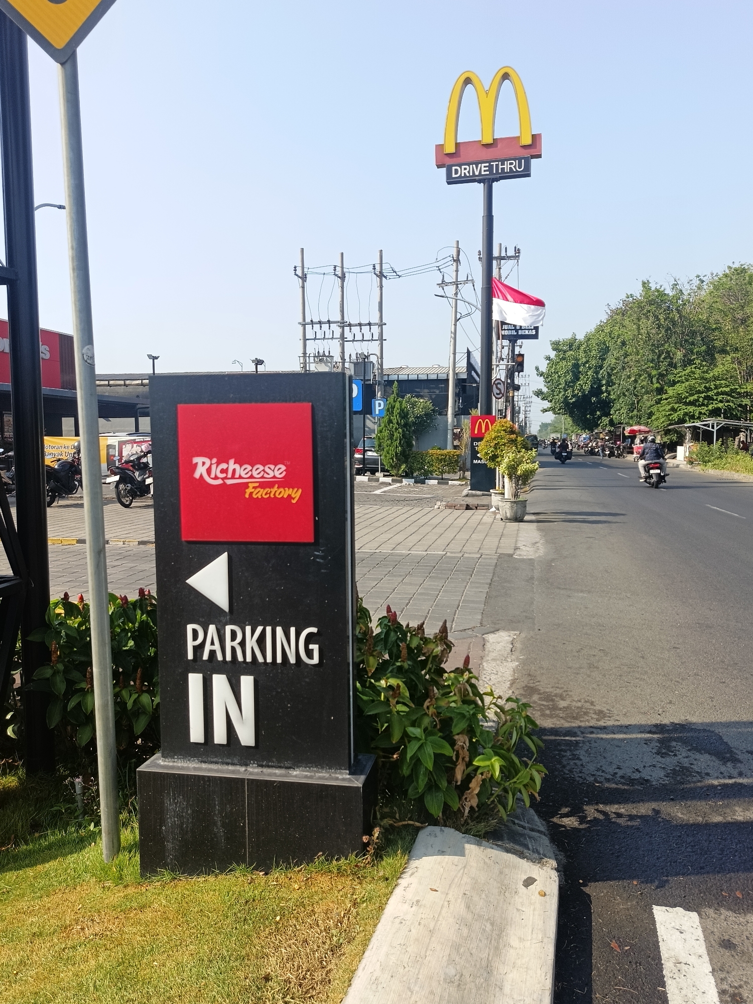 The image depicts a street scene in Calukan RT 1 RW 5 Keboansikep, Indonesia, on a clear day. In the foreground, a black sign with white lettering indicates "PARKING IN" and features a red square with the "Richeese Factory" logo. To the left of this sign, a tall gray pole with a yellow triangular sign is visible. Further down the street, a McDonald's Drive-Thru sign stands atop a pole, with an Indonesian flag flying nearby. The road is paved, with several motorcycles and a few cars present. Trees line the right side of the road, and some buildings are visible in the background. The overall scene suggests a commercial area.
