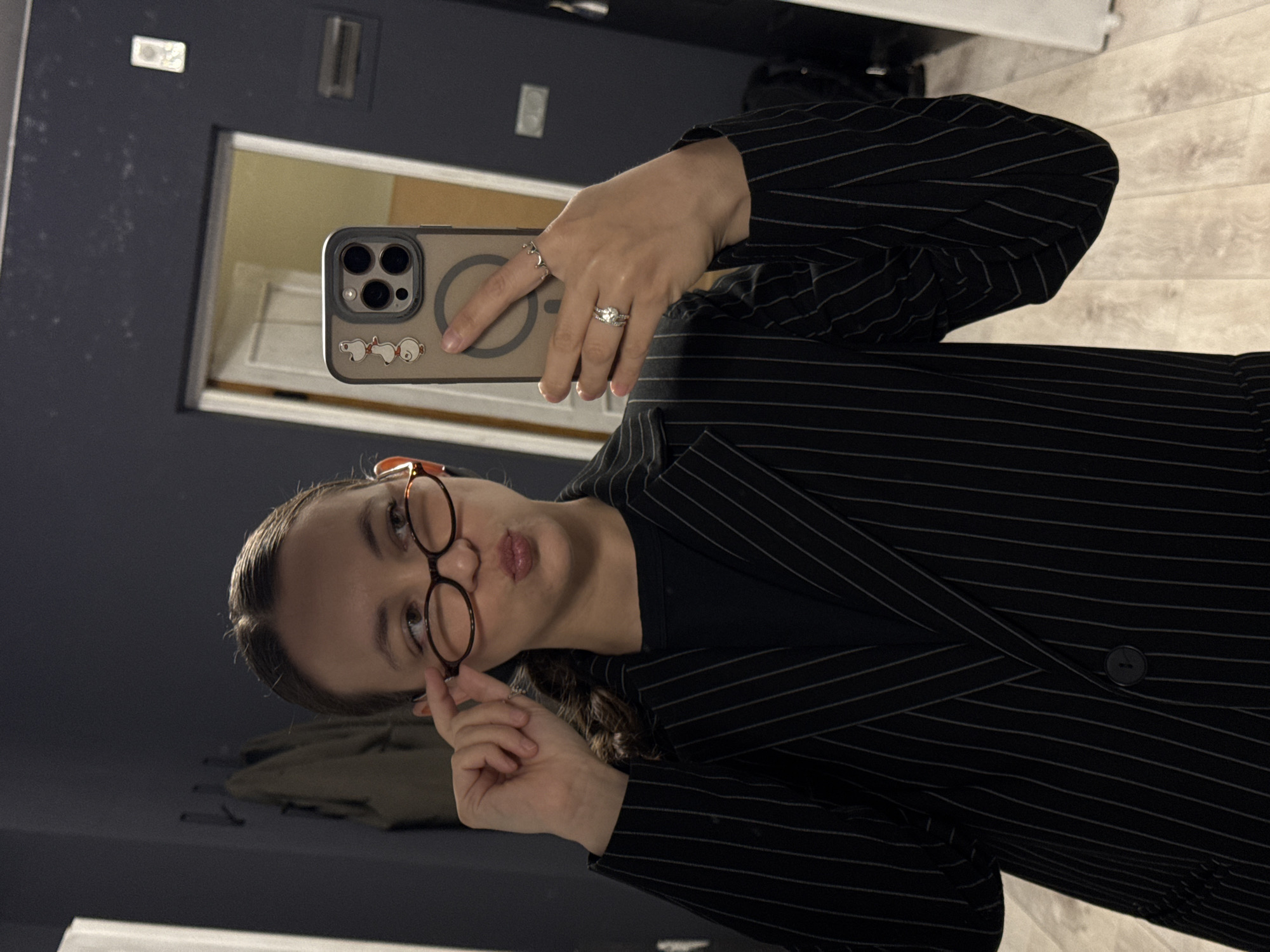In this media file, a young woman is seen taking a selfie in what appears to be a dimly lit room, possibly a bathroom or dressing area, given the presence of a mirror and a wall-mounted fixture. The woman is wearing a black pinstripe blazer over a black top. She has on round, tortoiseshell-framed glasses and is holding her smartphone in her left hand, which is angled towards the mirror. Her right hand is raised, with her index finger touching the edge of her glasses, and her lips are pursed. She is also wearing several rings on her fingers. The smartphone case is a light beige color and features a circular magnetic charging area, as well as small cartoonish decals of white figures. The background shows a reflective surface, likely a mirror, revealing a closet with hanging clothes and a wooden door. The lighting is artificial and somewhat subdued, suggesting it could be evening or a room with limited natural light. There is no visible text in the image.