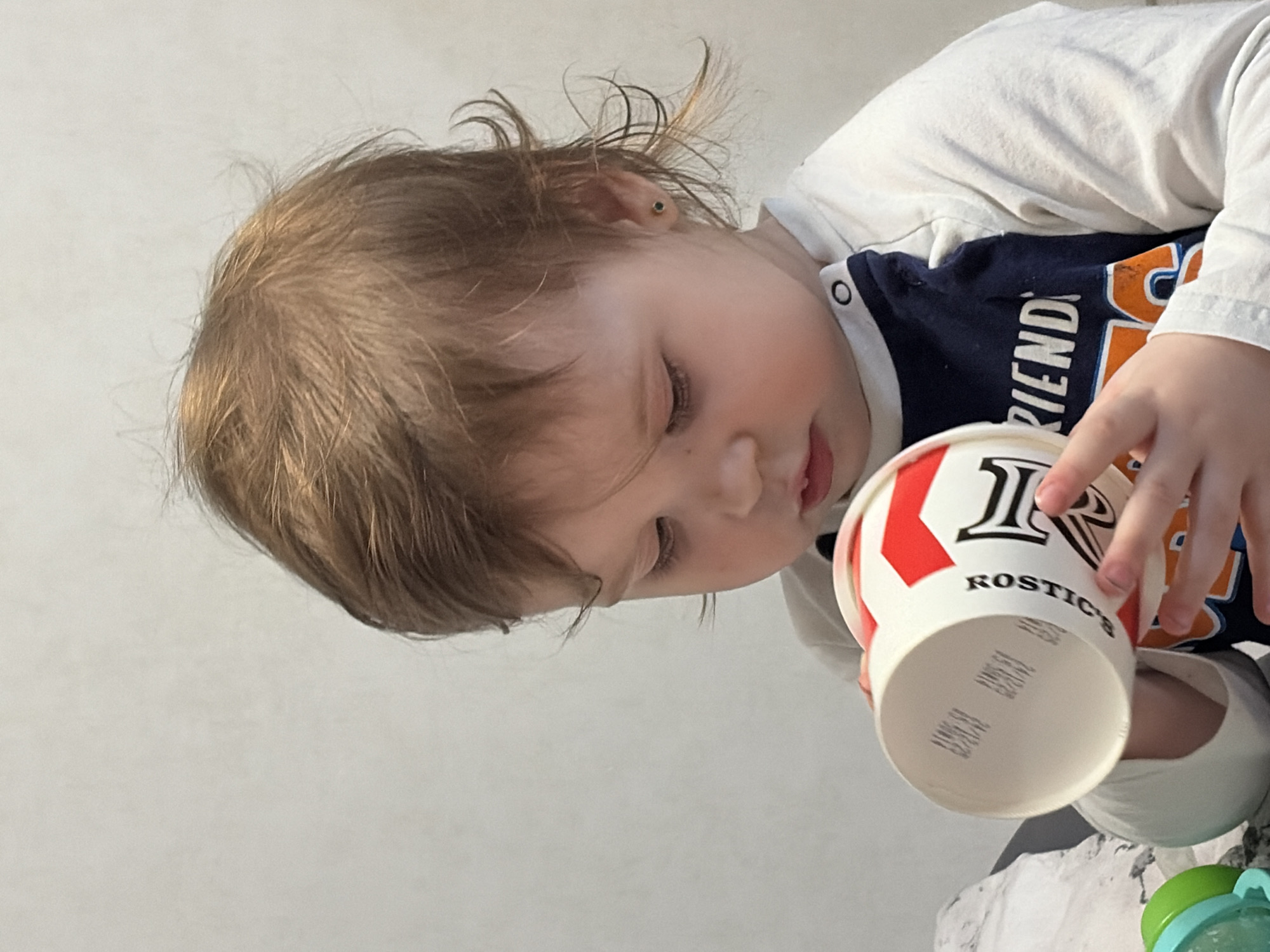 This image, taken in Chaikovsky, Russia, depicts a young child in a close-up, chest-up view against a plain, light-colored wall. The child has light brown hair and fair skin, with a small, dark stud earring visible in their right ear. They are wearing a long-sleeved, raglan-style shirt with white sleeves and a dark blue body, featuring white text, partially visible as "FRIEND". The child is holding an inverted white paper cup with both hands, looking down into its opening. The cup displays red and black graphics, including the word "ROSTIC'S" printed vertically along its side. The bottom of the cup shows two rows of indistinct imprinted or embossed characters. In the lower-left foreground, a partially visible green and blue plastic object rests on a light-colored, patterned fabric surface.