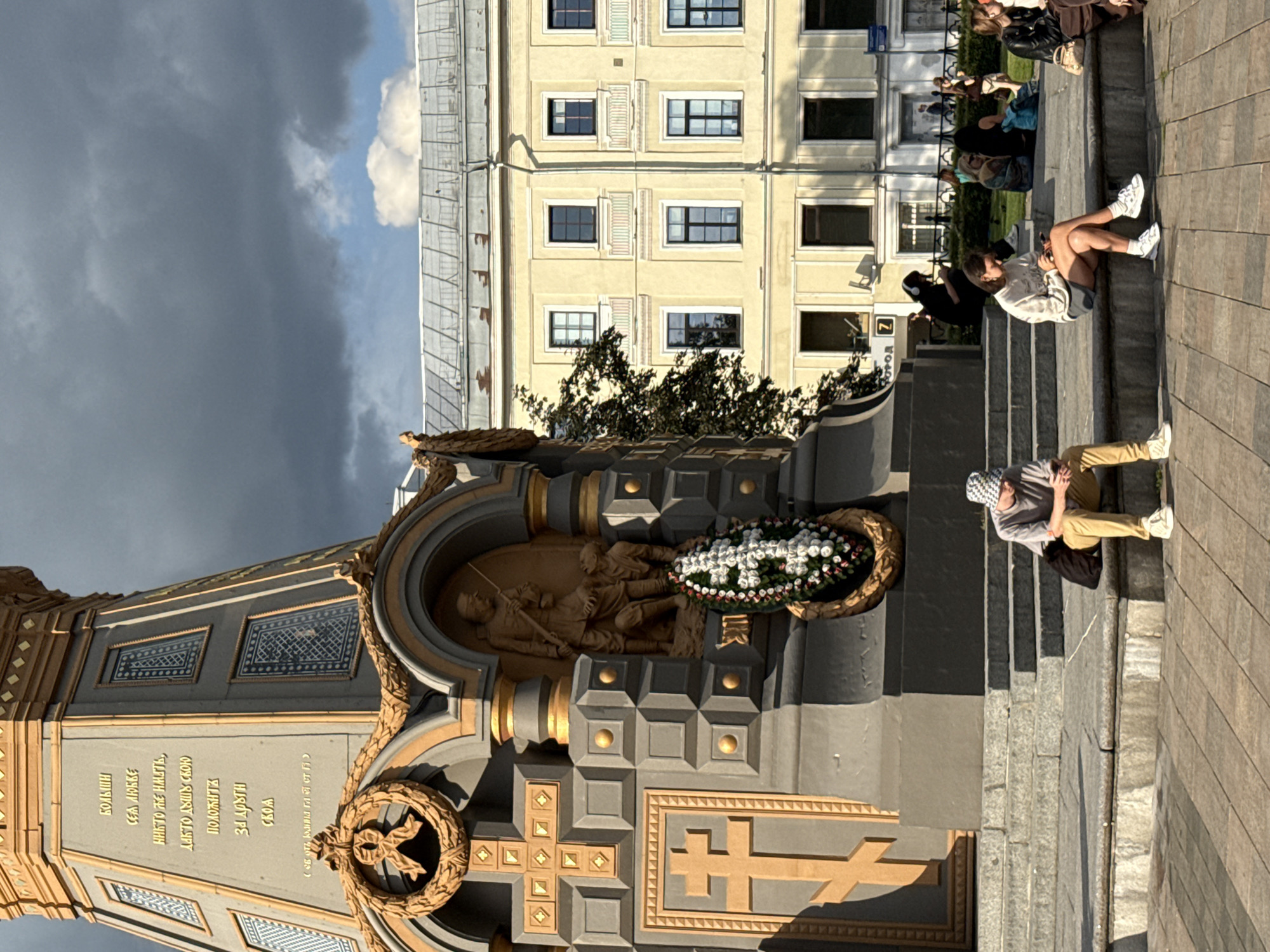 The image shows a monument with inscriptions in Russian. There are several people sitting on the steps of the monument, some looking at their phones. In the background, there is a yellow building with multiple windows. The sky is cloudy. This monument is located in Moscow, Russia, at the monument to Alexander Suvorov.