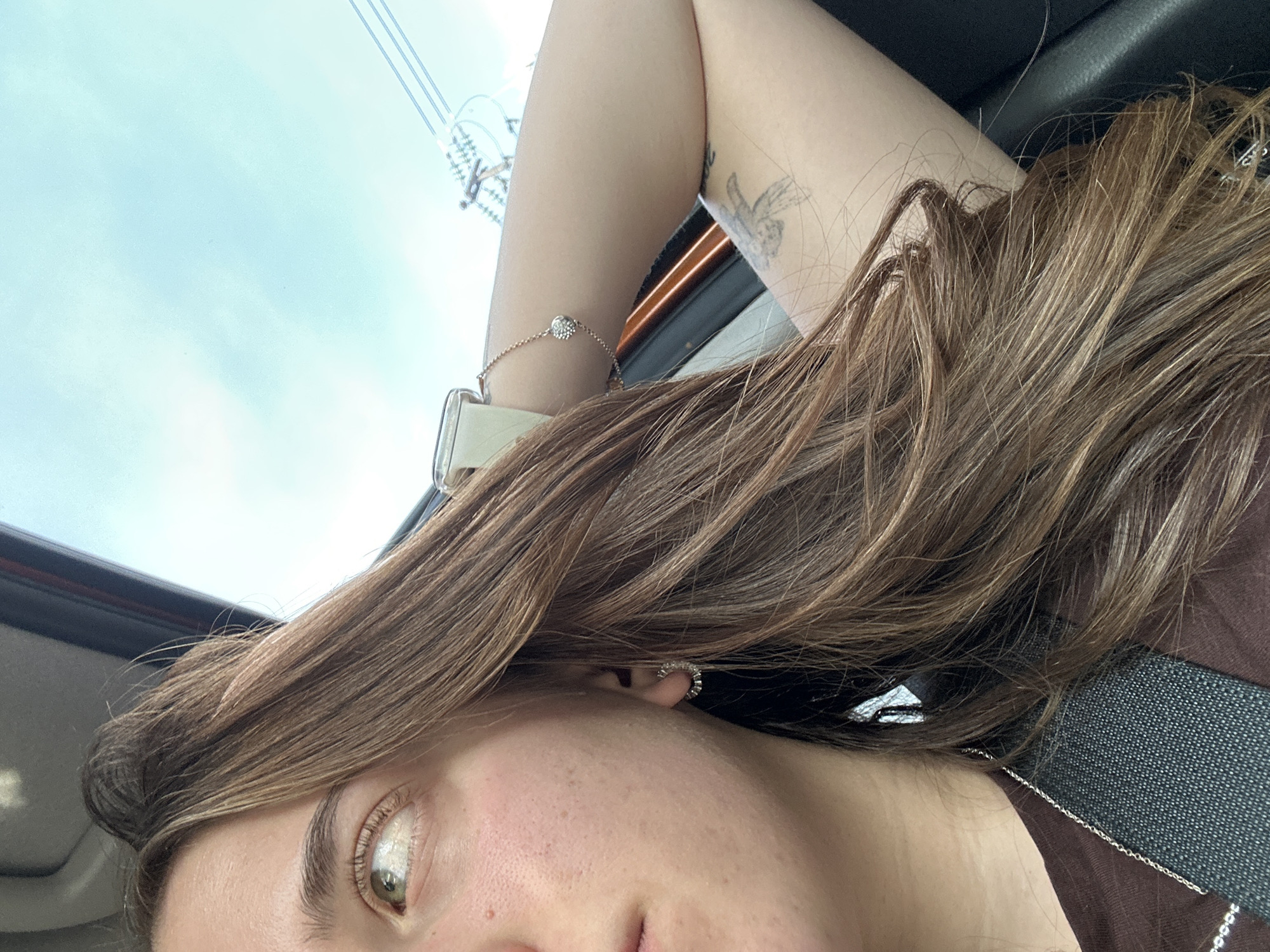 A woman is seated in a car, as evidenced by the safety belt visible across her brown shirt. Her left arm is raised, displaying a tattoo on her inner arm, a bracelet, and a watch on her wrist. The exterior view includes power lines and the sky. It is not possible to verify the location as Huixquilucan de Degollado, Mexico, based on the provided image.