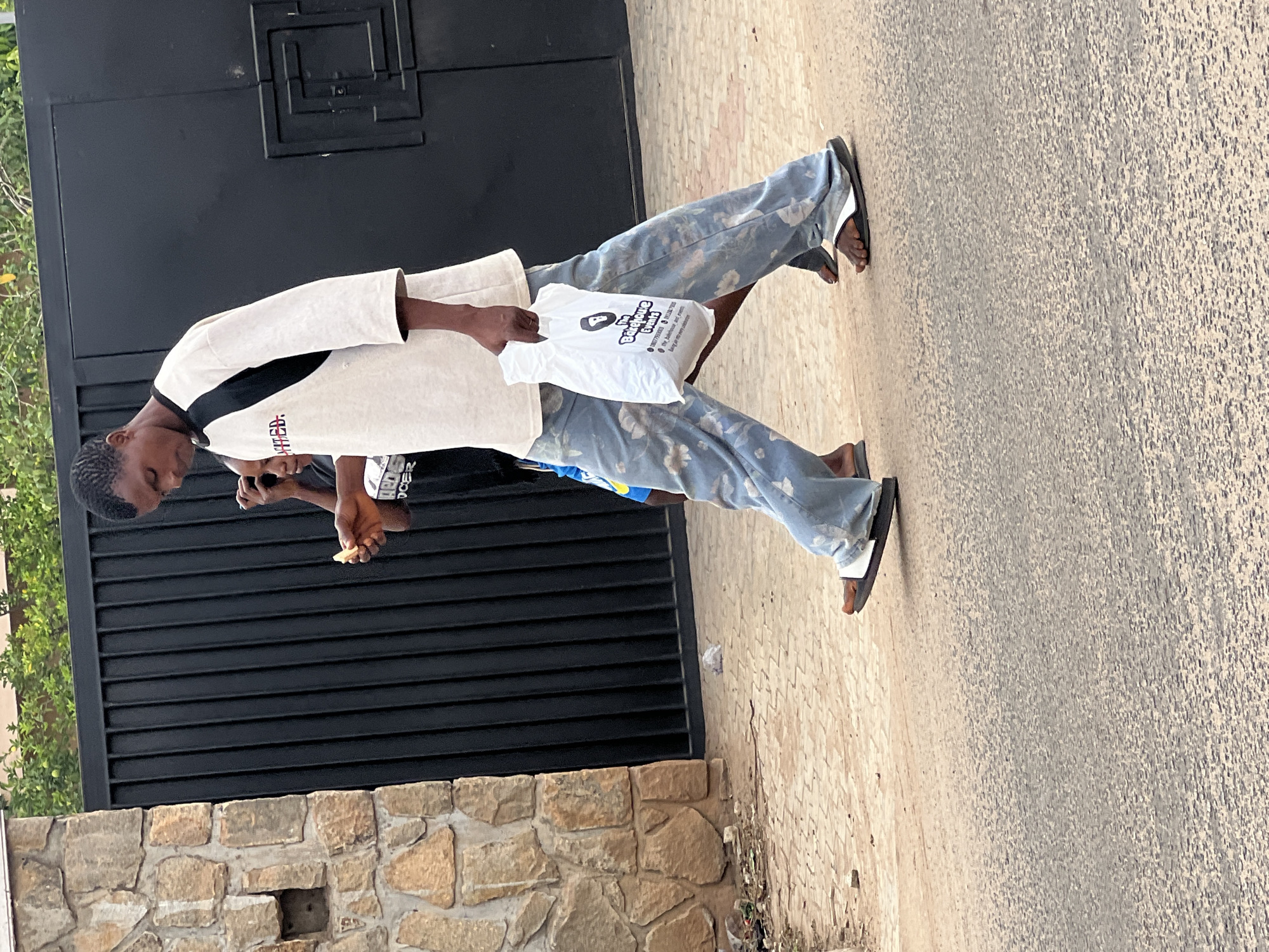 The image displays an outdoor scene in Gura Top, Nigeria, featuring two individuals. The central figure is a young person, likely male, wearing a light-colored (possibly off-white) long-sleeved shirt with a black section on the left shoulder/arm and light blue trousers with white and light blue cloud-like patterns. This individual is wearing dark flip-flops and holds a white plastic bag in their right hand. Partially visible red text is present on the chest of their shirt.
A second person is partially obscured behind the primary subject, wearing a dark top with unreadable white text and light blue shorts or pants. This individual appears to be holding a small, light-colored piece of food.
The background consists of a tall, dark, possibly metallic gate or wall with a decorative geometric pattern at its top. Beneath this gate is a rough-hewn stone wall made of varying shades of brown and beige blocks. To the far left, green foliage is visible. The ground is a paved surface, showing different textures, from lighter paving stones near the individuals to a darker, more textured road surface further away.
The white plastic bag held by the main subject prominently features the text "Babyblue events" along with a logo depicting a stylized head silhouette. Below this, contact details including "+234" (Nigeria's country code) and a phone number are visible. Services like "birthdays," "weddings," "engagements," "surprise events," "private dinners," and "party planning" are also listed, indicating an event management company.
The scene is well-lit by natural light, suggesting it is daytime. The overall impression is that of a casual, everyday street or roadside interaction.