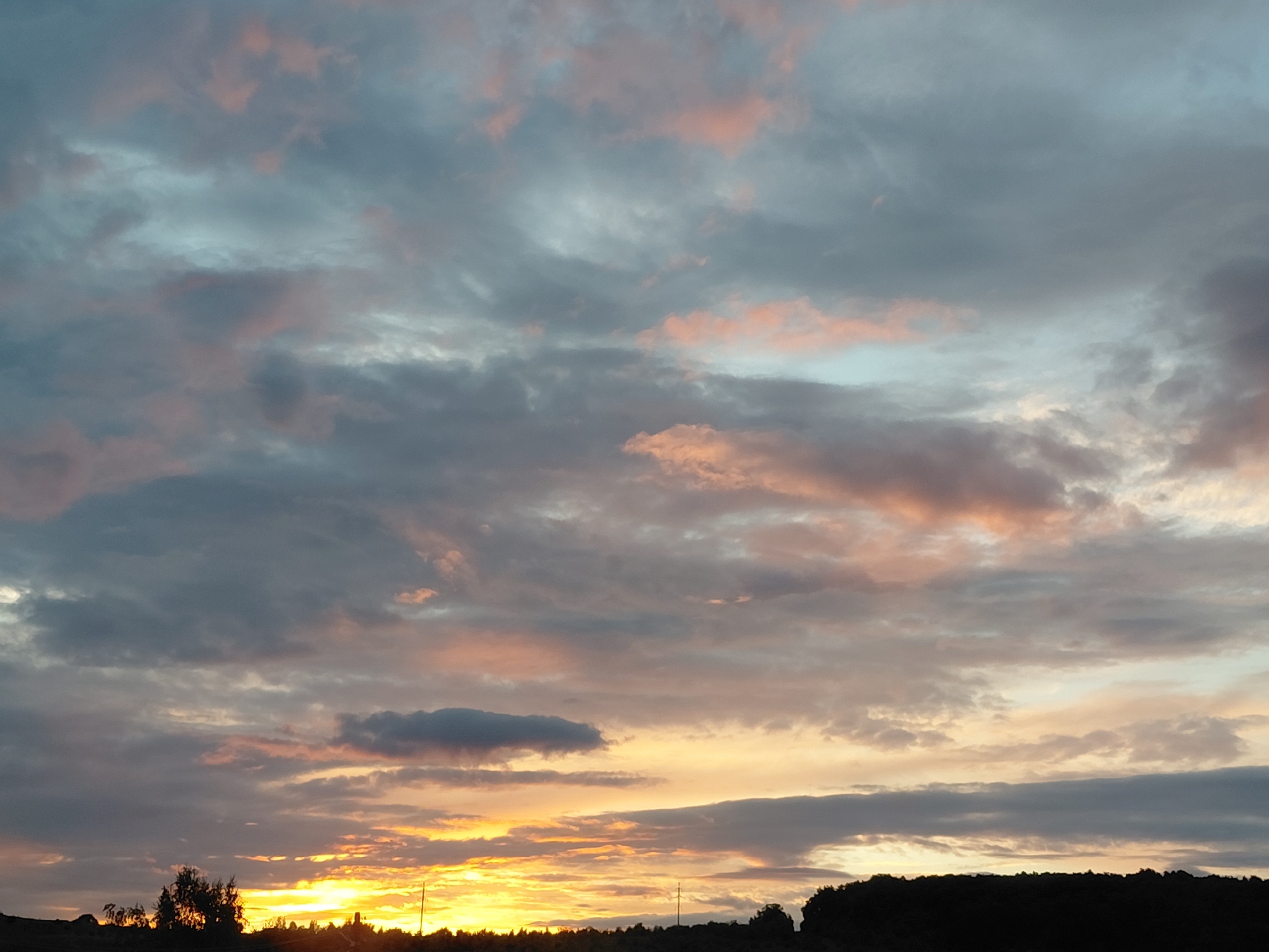A dramatic sunset scene unfolds over a silhouetted landscape in Novokuybyshevsk, Russia. The sky is a canvas of heavy, textured clouds, painted with hues of grey, soft pink, and pale blue. Wisps of darker grey clouds are interspersed with lighter, more diffused formations that catch the warm glow of the setting sun. The sun itself is not directly visible, but its intense golden-orange light emanates from behind a band of low-lying clouds near the horizon, casting a vibrant glow that illuminates the undersides of the clouds above and creates a bright, luminous layer across the lower sky.
The foreground is dominated by a dark, silhouetted treeline, suggesting a rural or park-like setting on the outskirts of the city. The shapes of trees and possibly some shrubbery are indistinct against the bright sky. A few vertical poles, likely utility poles or streetlights, are faintly visible as thin dark lines against the horizon. The overall atmosphere is one of natural beauty and the concluding moments of daylight. The scene appears to be devoid of any human figures or active subjects.