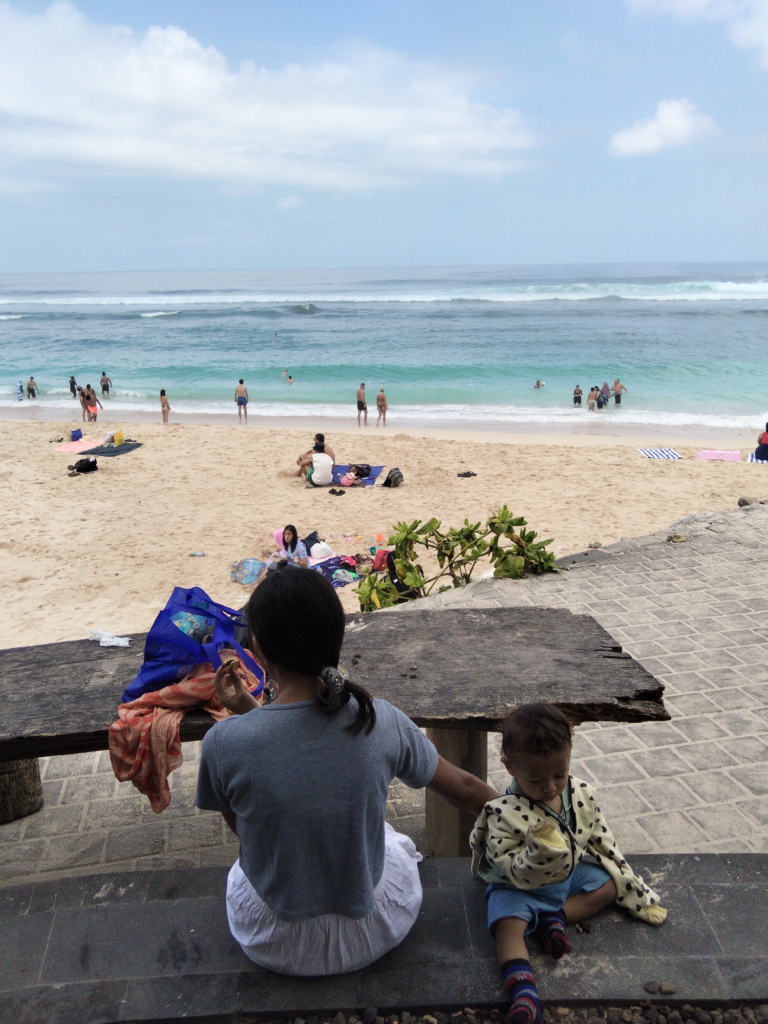 In Kuta Selatan, Indonesia, a beach scene unfolds under a partly cloudy sky. The ocean, with its distinct turquoise hue, features gentle waves rolling towards a sandy shore. Several individuals are visible in the water, some wading, others swimming. Along the beach, people are scattered, some sitting on towels, others standing or walking.
In the foreground, two children are seated on a dark-colored surface, possibly a concrete or stone wall, facing away from the camera. A young girl, positioned to the left, wears a grey shirt and a white skirt, with a blue bag and some fabric items on a wooden table beside her. To her right, a younger child, likely a boy, is dressed in a light-colored jacket with black polka dots over blue shorts and striped socks. He is looking downwards, holding a yellow object. The wooden table behind them appears weathered and aged. The background showcases the expanse of the beach and the ocean.