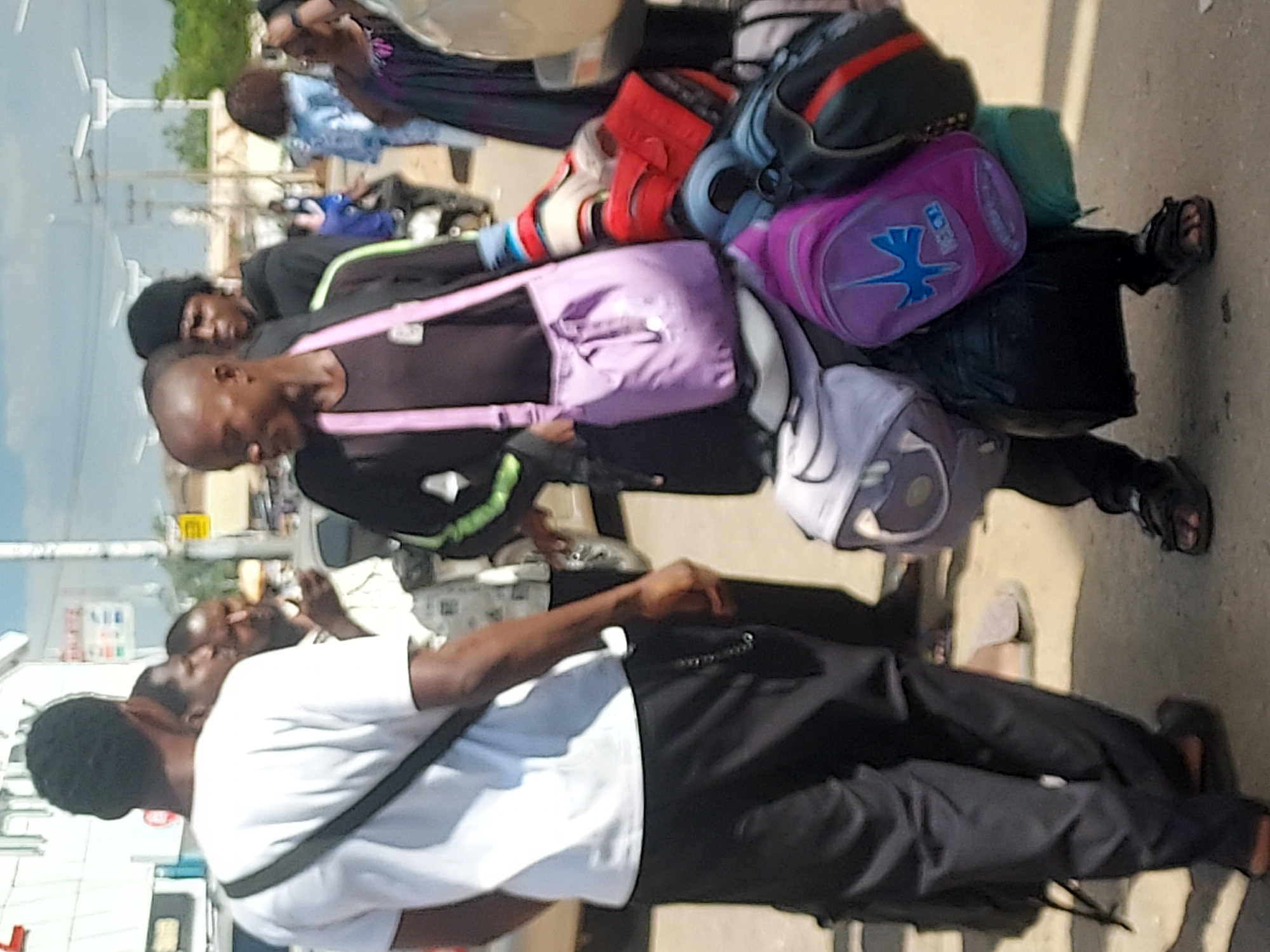 The image, taken in what appears to be a bustling outdoor public space in Jos, Nigeria, during daylight hours, shows several individuals, many laden with multiple bags.
In the foreground, a man with dark skin and close-cropped dark hair is seen from the back and side, wearing a white t-shirt, black pants, and a dark strap across his chest. He faces towards the right of the frame. To his left, another man, also with dark skin, is heavily burdened with luggage. He wears a dark shirt with a light green stripe and carries a variety of bags, including a prominent purple shoulder bag and a bright pink duffel bag. The pink duffel bag features a blue logo, possibly an abstract design or a stylized bird, and some faint text that might be "TOGA". Among his other belongings are a grey backpack, a black bag with red accents, and a distinctive red and white item that resembles a sports accessory or a boxing glove. A light green bundle is also visible among his load.
Several other individuals are present in the midground and background, some also carrying bags. One person further back is seen with a purple handbag. The setting appears to be a busy thoroughfare or transit point, with poles, distant structures, and some sparse vegetation in the background. The ground is a light, dusty color, possibly indicating a dirt road or paved area. The bright lighting suggests a sunny day. The scene captures a moment of everyday activity, likely related to travel, commuting, or the transport of goods, with people moving through the space. No strong emotions are evident on the visible faces.